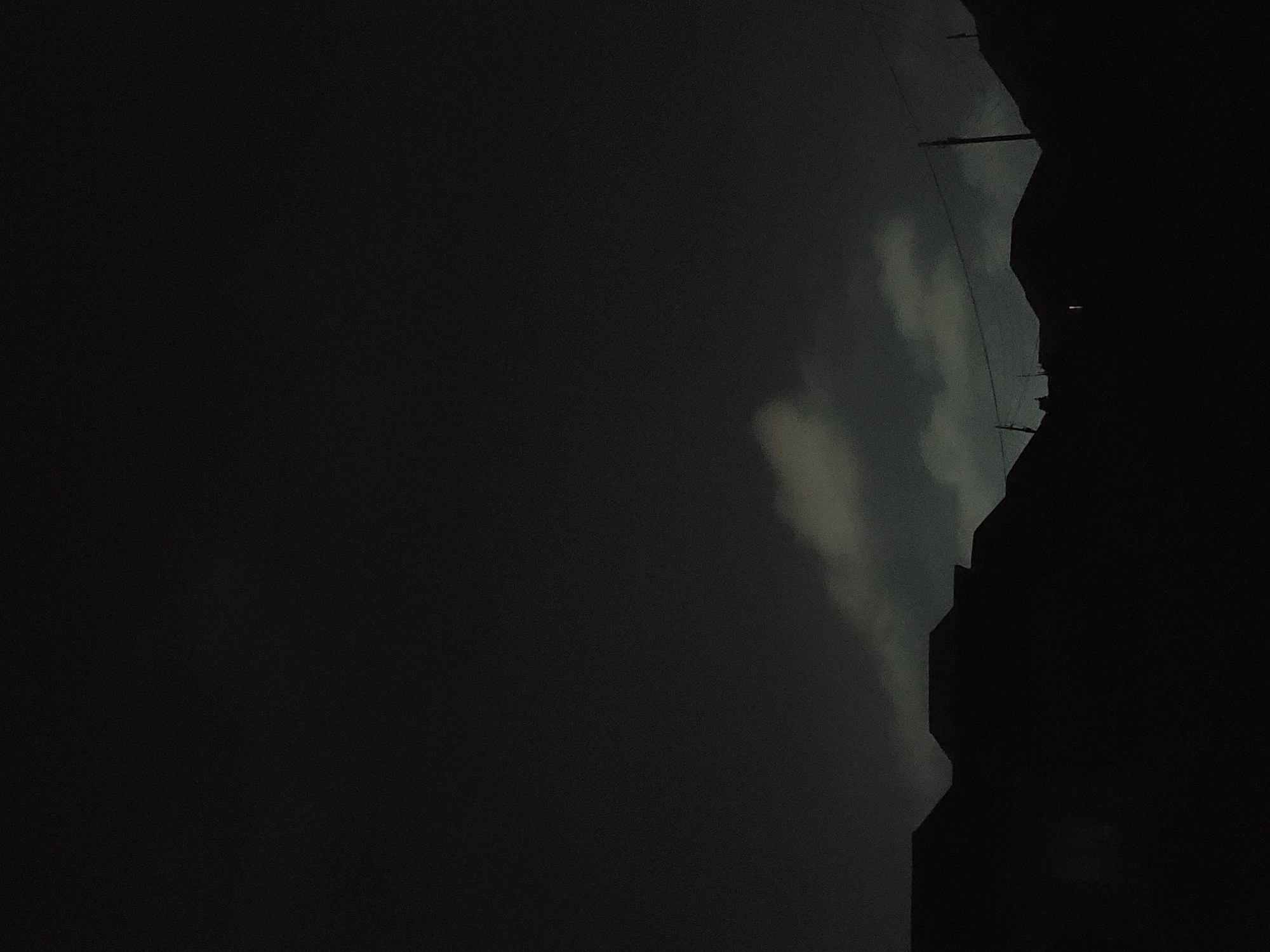 This image captures a nighttime scene in Jos, Nigeria, dominated by a dark, cloudy sky. Silhouetted against the sky are the rooftops of several houses and buildings, creating a stark outline of an urban landscape. The clouds appear somewhat illuminated, suggesting the presence of ambient light, possibly from the moon or distant city lights, though no distinct light sources are visible. A few utility poles with wires are also discernible in silhouette, indicating the presence of infrastructure. The overall impression is one of a quiet, subdued evening in the city.