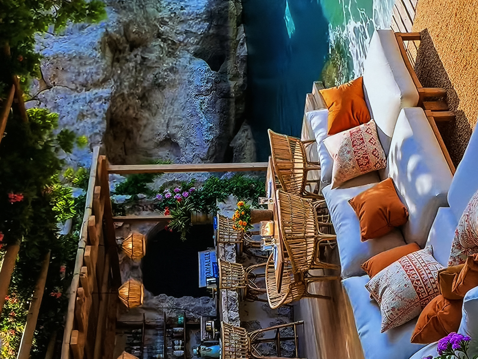 The image displays an inviting outdoor lounge or restaurant area situated by rocky cliffs and clear blue-green water. The scene is presented from an elevated perspective and is rotated 90 degrees counter-clockwise.
In the foreground, a wooden deck holds comfortable seating arrangements, including white padded benches and sofas generously accessorized with throw pillows in burnt orange, terracotta, and intricate patterned designs. Wicker chairs and wooden tables are interspersed throughout the area, complemented by hanging wicker lanterns, some of which are illuminated, casting a warm glow. Potted plants with vibrant pink, purple, and orange flowers add decorative touches.
To the left, imposing natural rock formations rise, characterized by rugged textures and shades of gray and blue. Lush green foliage and tree branches partially frame this side of the scene. Between the seating area and the cliffs, a calm body of water shimmers with clear blue-green tones, suggesting a tranquil coastal or lakeside setting.
A bar or service counter, constructed from stone and wood, is visible under a wooden pergola-like structure. Above this counter, a blue sign displays text that reads "PRAYALUZ". The ambient lighting, provided by the lanterns, suggests an evening or late afternoon time of day.
The overall atmosphere is one of serene relaxation and natural beauty, characteristic of a luxurious resort or beach club. No people are visible. While the visual elements strongly suggest a coastal environment, the provided location context is Sofiivska Borshchahivka, Ukraine.