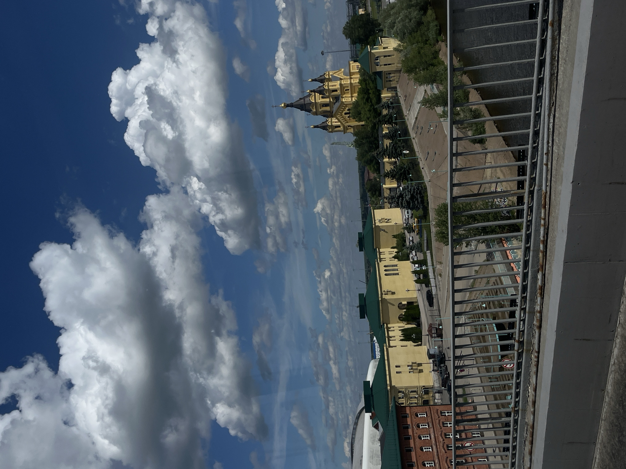 The image shows an aerial view of a city, featuring a large yellow cathedral with multiple domes, a waterfront, and several buildings with green roofs. The sky is blue with fluffy white clouds. In the foreground, a metal railing is visible, suggesting the photo was taken from a bridge or elevated walkway. There are cars parked along a road, and trees line the waterfront. The overall scene depicts a sunny day in a European-style urban setting.