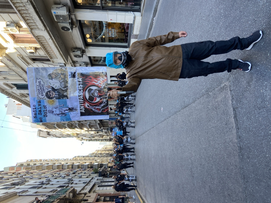 A lone individual stands on an asphalt street in Buenos Aires, Argentina, facing slightly right of the viewer's perspective. The person, a male in a brown leather jacket, black cargo pants, and black sneakers, wears a light blue head covering and a black full-face gas mask. His right arm is raised, holding a rectangular protest sign aloft. The sign's upper section displays a blue and white background with two panels: one depicting a figure with "Nadie se Salva Solo!" (No one saves themselves alone!), and another with a caricature of a man in a suit proclaiming "¡VIVA LA ESCLAVITUD, CARAJO!" (Long live slavery, damn it!) and "12 hs" alongside a worker. The sign's lower section is black, featuring a red graphic of a hooded figure holding a flaming torch, overlaid with the white text "ANTIFASCISTA."
In the midground, a line of approximately 10-15 uniformed police officers, wearing helmets and dark uniforms, form a barrier across the street. Each officer holds a transparent riot shield, several marked with "POLICIA." A light blue police vehicle is partially visible behind the police line. In the background, a crowd of people is gathered further down the street behind the police.
The street is narrow and lined on both sides by multi-story, aged residential and commercial buildings with numerous balconies and external air conditioning units. The sky is clear blue. One individual observes from a balcony on the far left. The architectural style, language on the sign and shields, and political context indicate the scene is in a central district of Buenos Aires, Argentina.