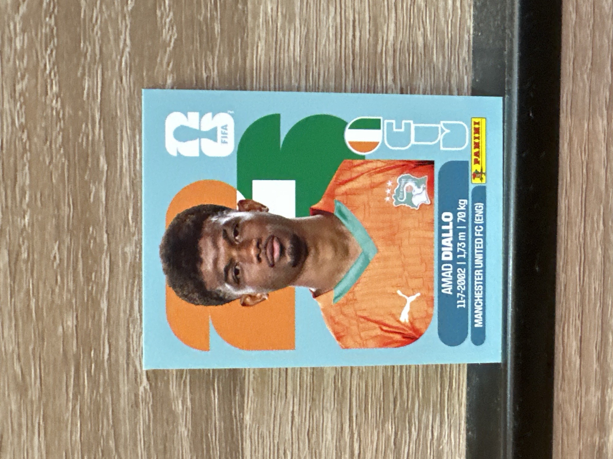 The image displays a Panini sticker of footballer Amad Diallo. The sticker has a light blue background with orange and green graphic elements. Amad Diallo is shown from the chest up, wearing an orange football jersey with a teal neckline. The jersey features a Puma logo and the crest of the Ivory Coast national team. Text on the sticker includes his name, date of birth (11-7-2002), height (1.73 m), weight (70 kg), and club (Manchester United FC (ENG)). A FIFA logo is also visible. The sticker is placed on a wooden surface, which appears to be a table or shelf. The background consists of a horizontal wood grain pattern. There is no indication of the city of Prisovo, Bulgaria, in the image or its context.