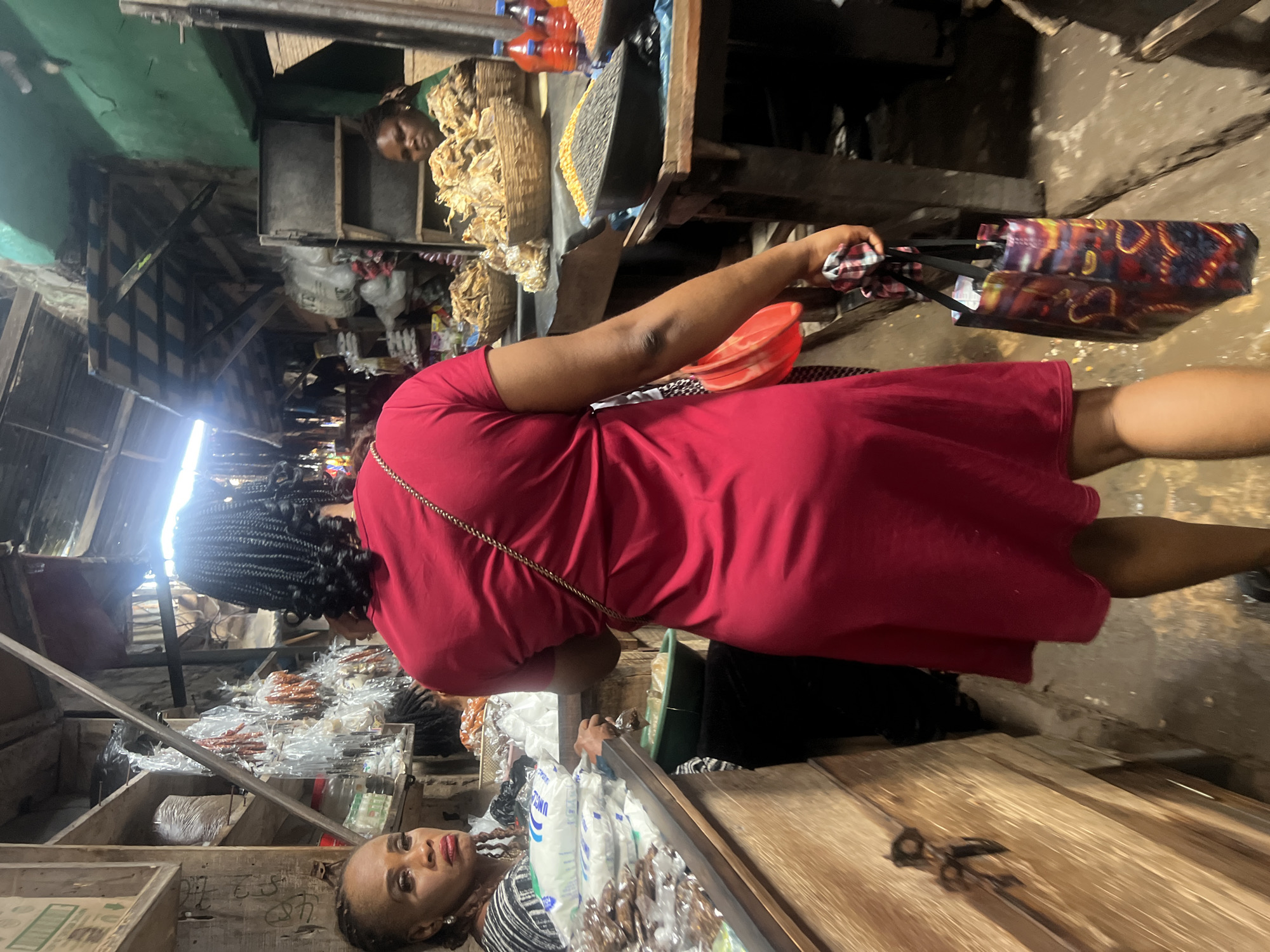 The image depicts a bustling market scene, likely in Nkpor, Nigeria. A woman in a red dress walks away from the camera, carrying a colorful reusable shopping bag. To her left, another woman, wearing a patterned shirt, looks towards the camera with a stern expression, her hands resting on a counter laden with bagged goods. Behind the woman in red, a stall displays various dried food items in woven baskets, and in the foreground, a black basin holds black-eyed peas. The market appears to be an open-air or partially covered structure with wooden stalls and a rough-textured floor. The overall atmosphere suggests a vibrant commercial setting with a variety of goods for sale.