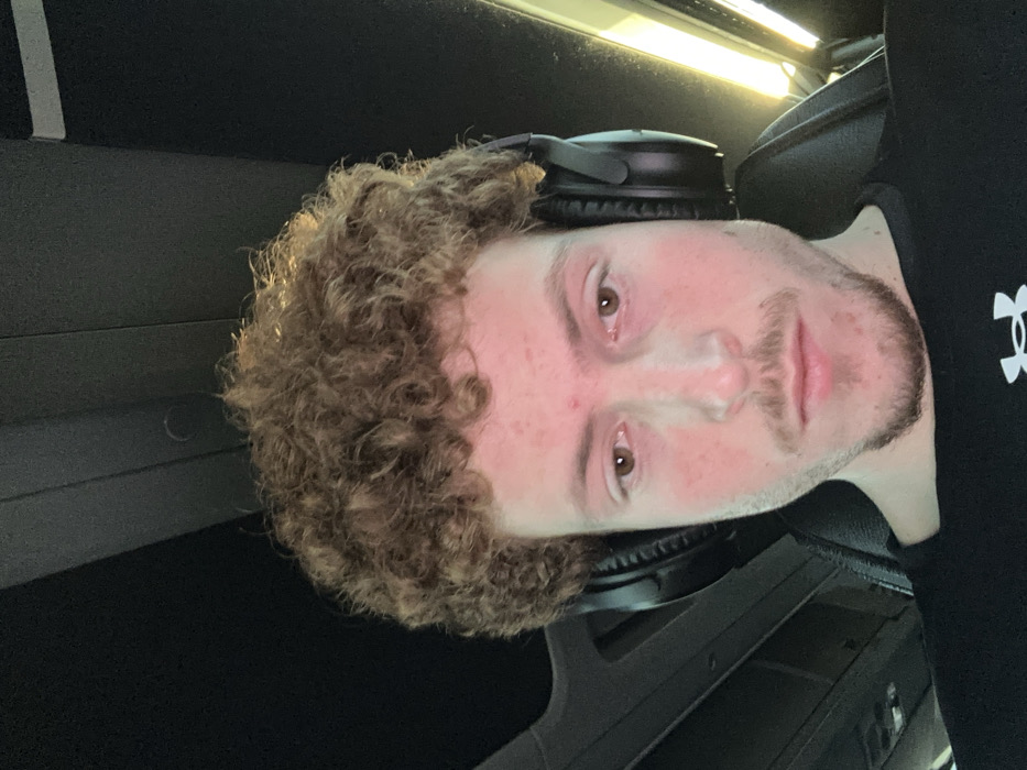 A fair-skinned person with curly auburn hair is wearing black headphones around their neck. The individual has light brown eyes, a stubbled beard, and a few blemishes on their face. They are wearing a black shirt with a visible logo on the right side. The background appears to be the interior of a vehicle, with dark panels and a light reflecting overhead. The image is rotated.