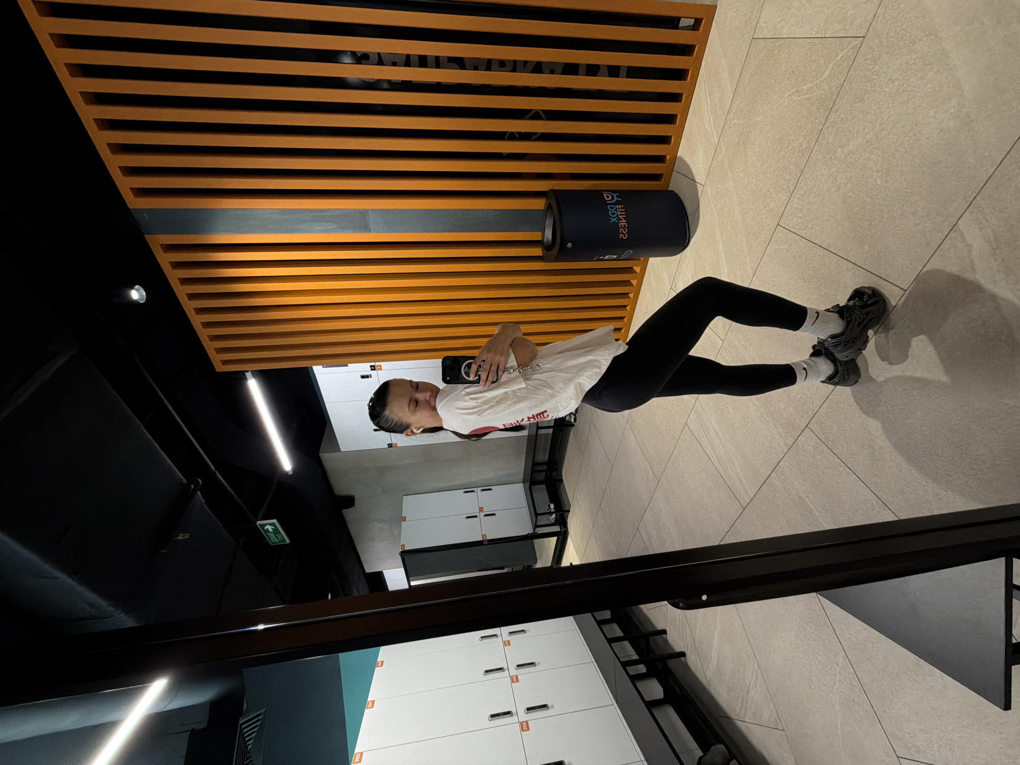 A young woman is standing in a gym locker room, taking a selfie with her phone. She is wearing black leggings, a white t-shirt, and gray sneakers. The locker room has white lockers and beige tile flooring. A ceiling fixture of orange and gray slats is visible above her. The gym is located in Moscow, Russia.