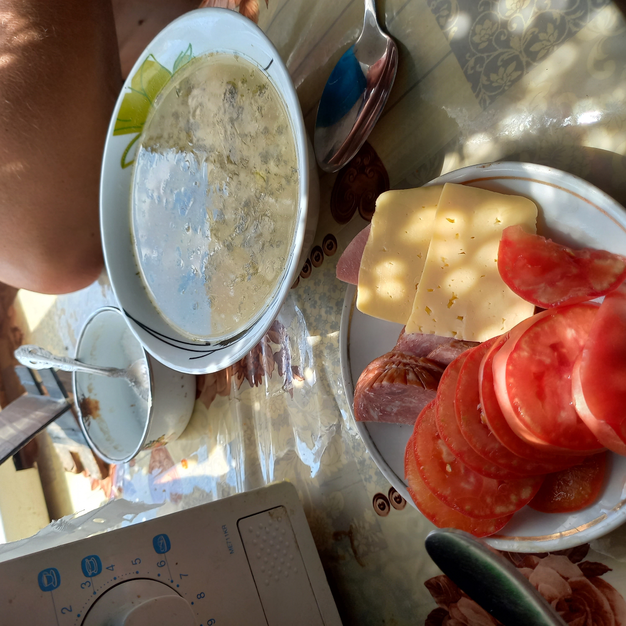 A close-up, eye-level shot captures a meal laid out on a patterned tablecloth. In the foreground, a white plate holds sliced tomatoes, chunks of sausage, and slices of yellow cheese. To the right, a large white bowl contains a light-colored soup with green flecks. A spoon rests in a smaller white bowl to the left of the soup. On the far left, a white appliance, possibly a sewing machine, is partially visible with a dial and buttons. A person's bare arm is visible in the upper right corner. The scene appears to be indoors, with natural light illuminating the food and objects. There are no discernible landmarks or context to suggest a specific location within Klyntsi, Ukraine.