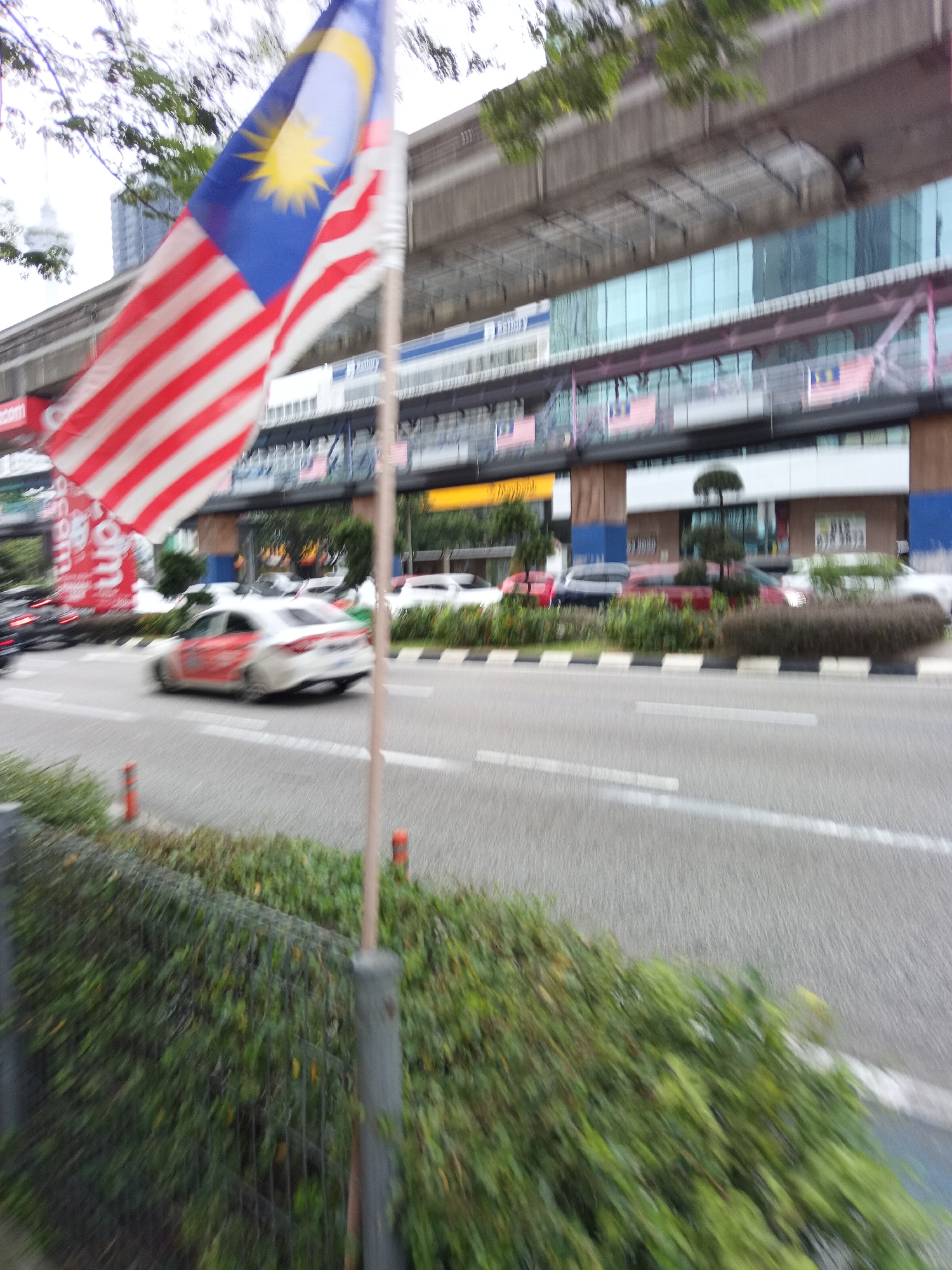 A blurry, outdoor, daytime, street-level shot captures a view of a busy road in Kuala Lumpur, Malaysia, with a Malaysian flag prominently displayed in the foreground. The flag, partially obscured by trees, is blowing in the wind and shows red and white stripes and a blue canton with a yellow crescent and star. In the middle ground, a white car with red accents is driving on the road, with other vehicles visible further down the street. Behind the road, a modern building with a glass facade and an elevated train track runs along the top. The surroundings include green foliage, a metal fence, and orange traffic cones. The overall impression is one of urban movement and national symbolism.