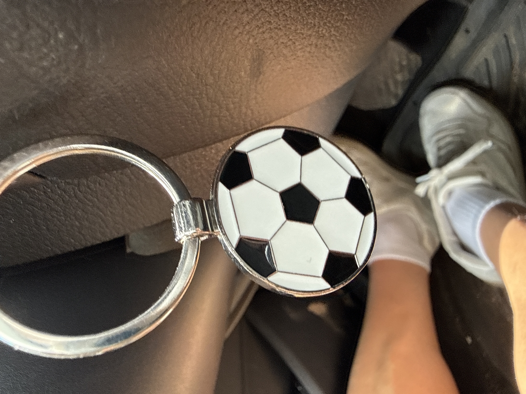 A close-up shot captures a shiny silver keychain featuring a circular charm with a classic black and white soccer ball design. The keychain's large ring is prominent on the left, while the soccer ball charm, depicting traditional hexagonal and pentagonal panels, is centrally placed and gleams under the light.
The scene appears to be set inside a car. A person's light-skinned leg and foot are visible in the lower right portion of the frame. The foot is clad in a white low-cut sock and a white lace-up sneaker, suggesting the person could be a passenger or a driver resting their foot.
The background consists of dark, textured surfaces, typical of a car interior, possibly parts of a seat, dashboard, or floor mat. Hints of car pedals can be discerned in the deeper background towards the bottom right, further confirming the car setting. The lighting indicates it is daytime. No visible text is present in the image. The location context for this image is Radinovo, Bulgaria.
