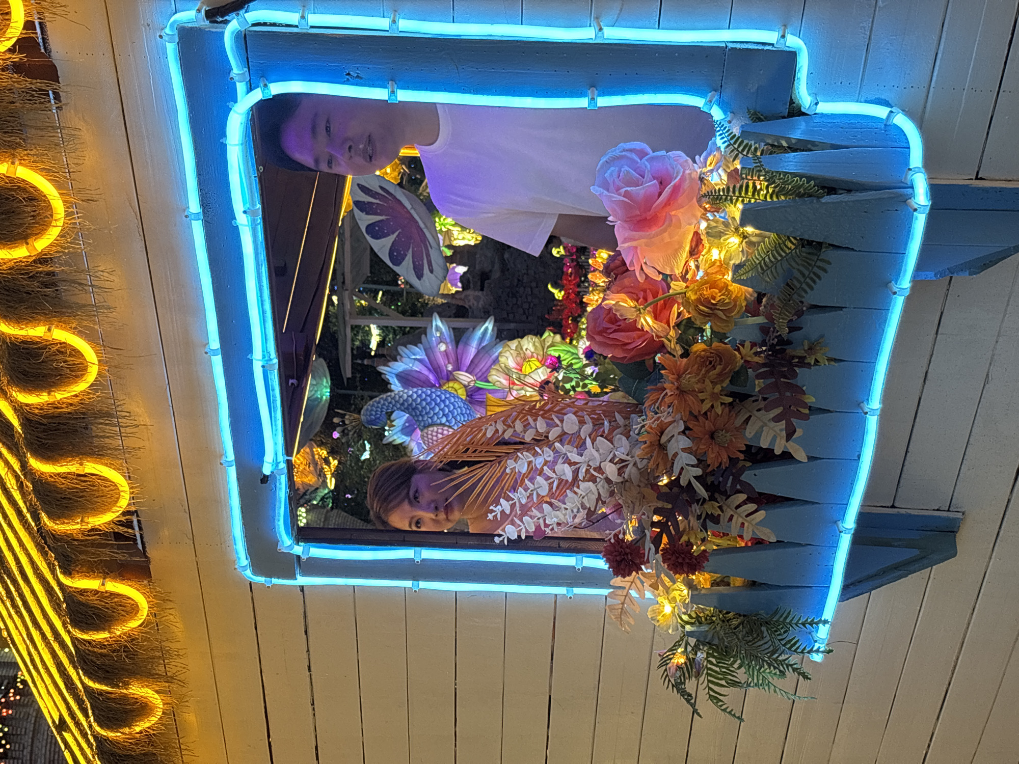 This image shows two people, a woman and a man, looking out of a window. The window is framed by bright blue neon lights. The woman is on the left, wearing a patterned dress. The man is on the right, wearing a white t-shirt. They appear to be in some kind of outdoor market or festival in Baqiao, China, as indicated by the festive lights and decorations visible in the background. The scene is illuminated by the blue neon lights and warm yellow lights from above. The window box in front of them is filled with artificial flowers and greenery.