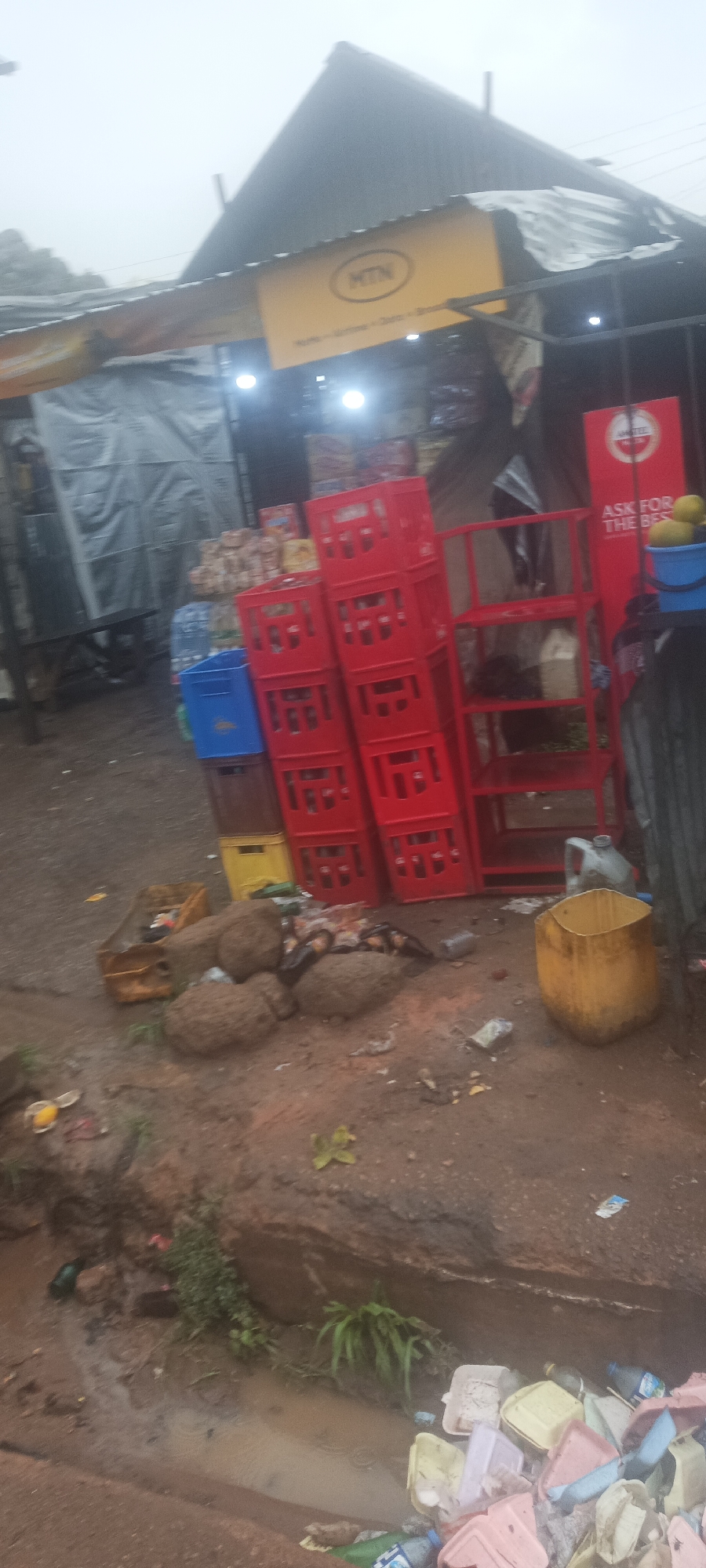 The image depicts a scene outside a store in Kuru Dungus, Nigeria. The store has an MTN sign and an Amstel beer advertisement. Stacks of red, blue, and yellow crates are near the store. In front of the store, there is trash. The ground is muddy.