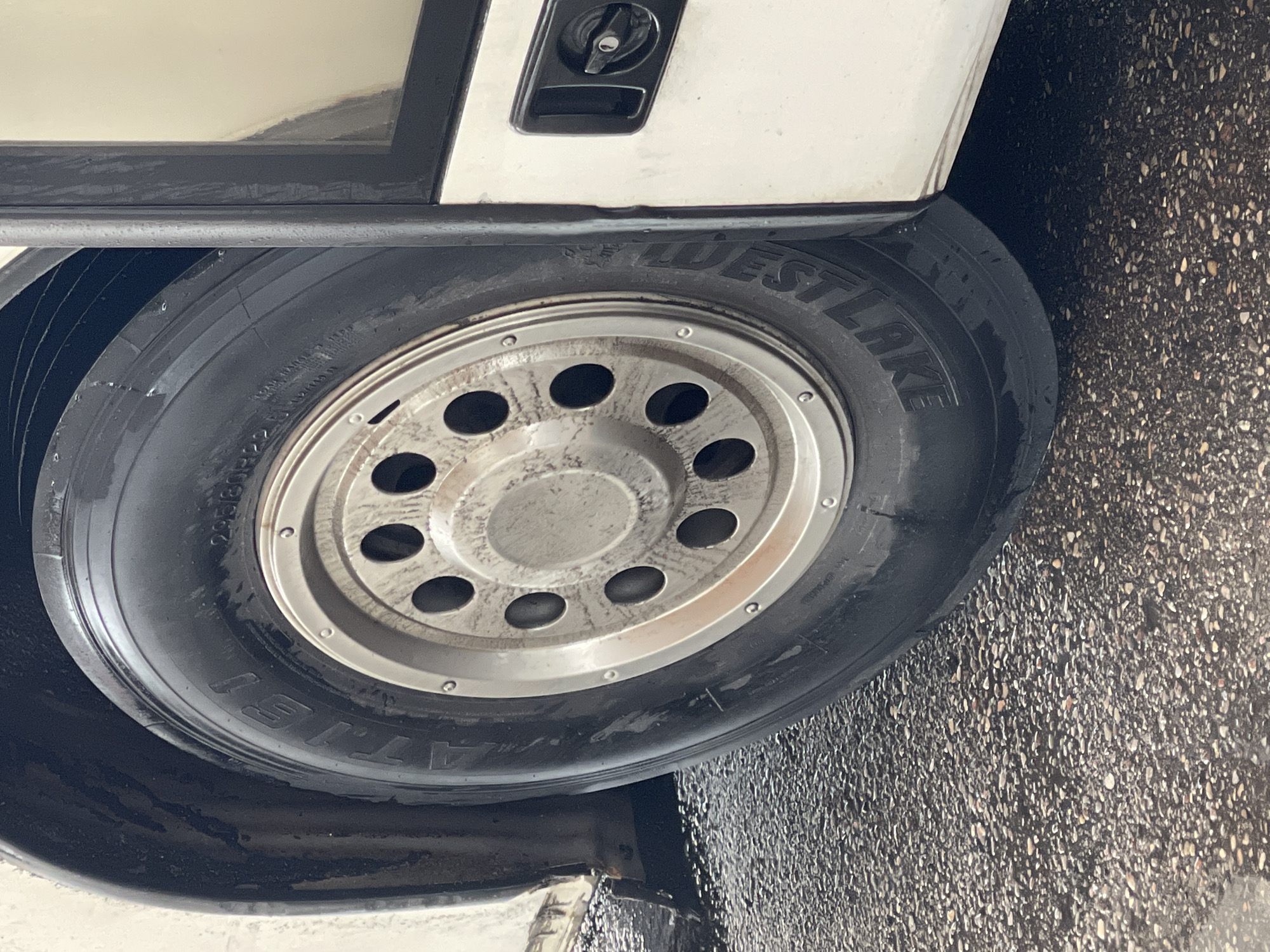 The image shows a close-up of a tire and wheel on the side of a white vehicle, possibly a bus or RV, parked on a wet, pebbled surface. The tire has the brand name "WESTLAKE" visible in raised lettering. The wheel is silver with multiple circular cutouts. The vehicle appears to be in an urban environment, but no specific landmarks in Zelenogradsk, Russia, are identifiable in the image. There are no people visible in the frame. The overall impression is of a stationary vehicle on a damp day, possibly after rain.