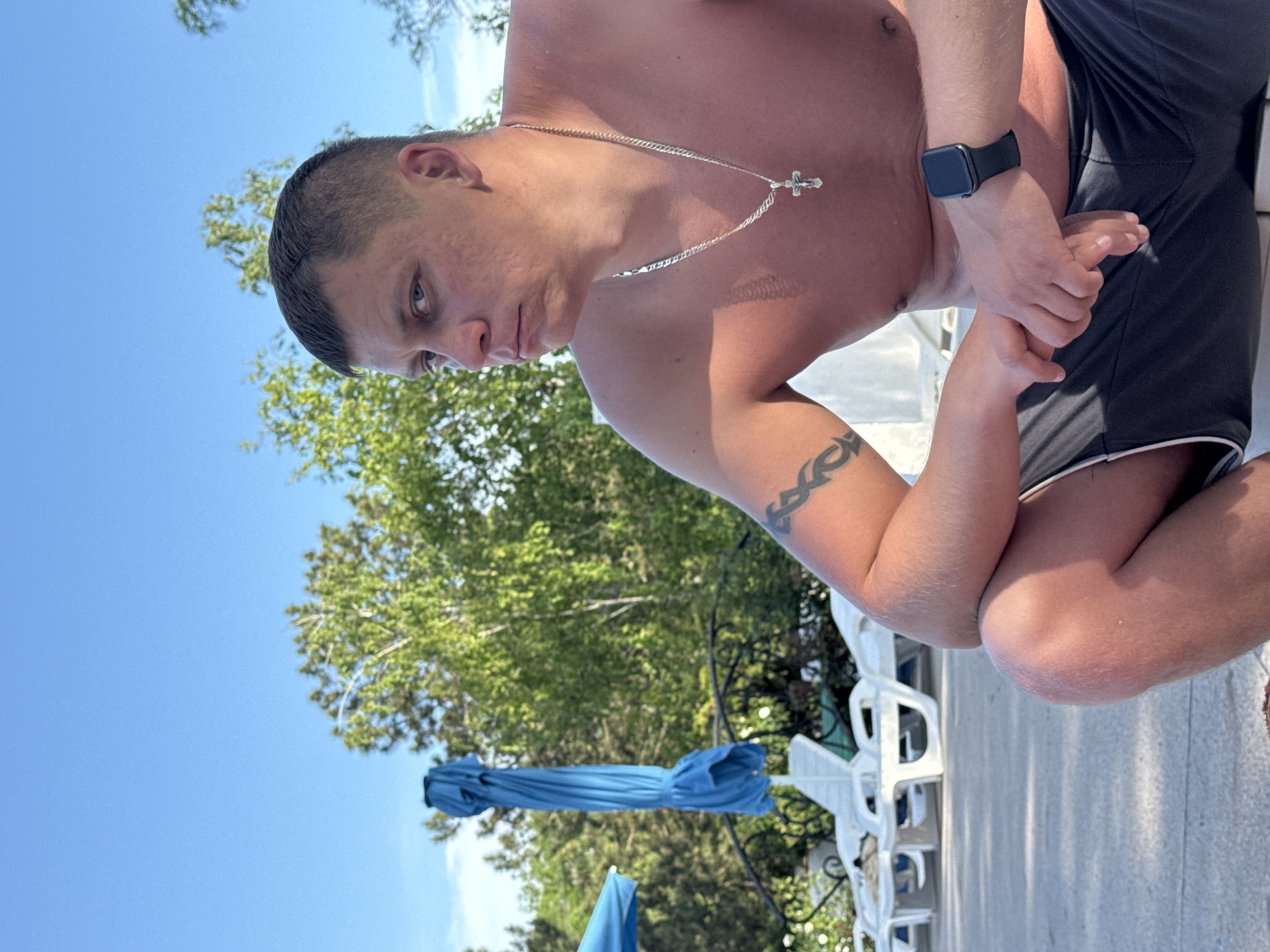 A man with short, dark hair and a cross necklace is crouched outdoors. He is wearing a black watch on his left wrist and has a tribal tattoo on his left arm. He is shirtless and wearing dark shorts with a white trim. Behind him, there is a blue sky, green trees, a closed blue umbrella, and white plastic chairs. The setting is likely a pool or leisure area. The exact location in Yevpatoriya, Ukraine, cannot be determined solely from this image.
