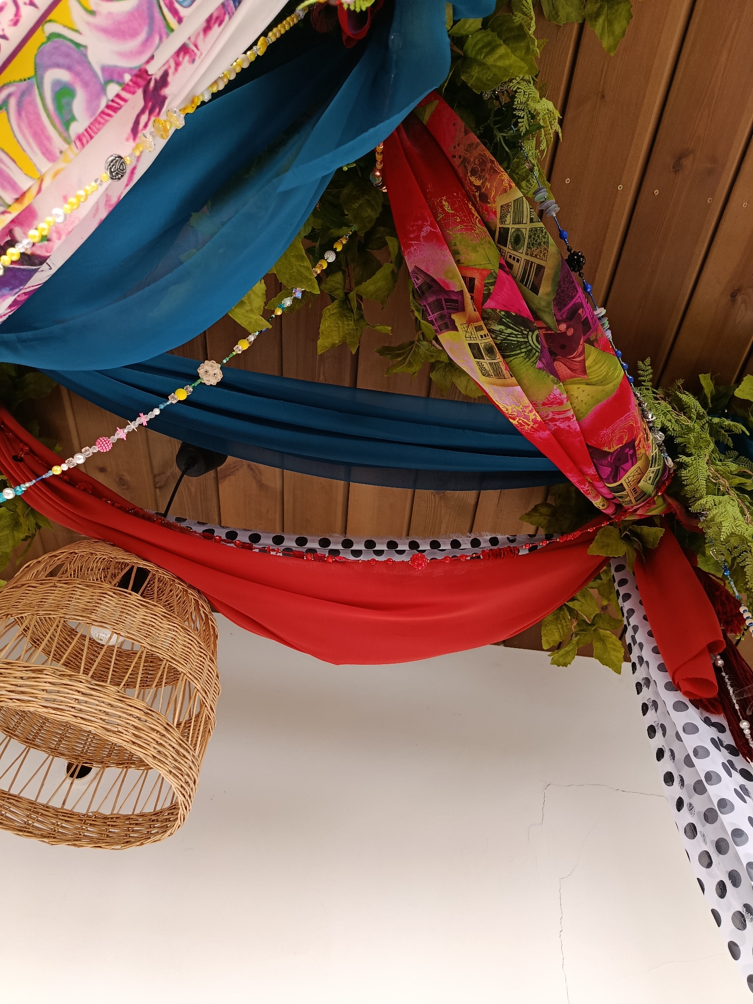 The image shows an overhead view of a decorated outdoor space. Several colorful fabrics, including a pink and white patterned fabric, a teal fabric, a red fabric, and a black and white polka dot fabric, are draped from a wooden ceiling. Green foliage is interspersed among the fabrics. Beaded strings are also hanging, some with colorful beads and charms. A woven wicker lampshade hangs on the left side of the frame, and a lightbulb is visible inside. The background shows wooden planks with visible screws. Below the draped fabrics, a white surface with some cracking is visible. The scene appears to be an exterior porch or patio decoration. There is no indication of the location being in Saratov, Russia.