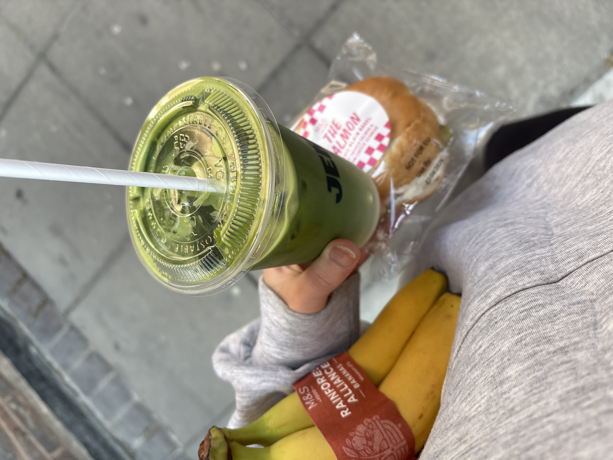 The image captures an individual carrying a collection of food items while navigating a gray, paved sidewalk. The individual's hand holds a green-colored beverage in a clear plastic cup with a white straw; the cup's lid is transparent and marked with the word "COMPOSTABLE." To the right of the drink is a plastic-wrapped bagel, with a label reading "THE SALMON." Below these items, several yellow bananas are visible, bundled together with a red band labeled "M&S RAINFOREST ALLIANCE - BANANAS." The person is wearing a gray sweater. The background consists of the sidewalk, which is a pattern of light and dark gray. The photo appears to have been taken from an above perspective, possibly by the person carrying the items. While it does not explicitly indicate that the location is London, United Kingdom, the M&S brand on the bananas is a clue, since it is a UK brand.