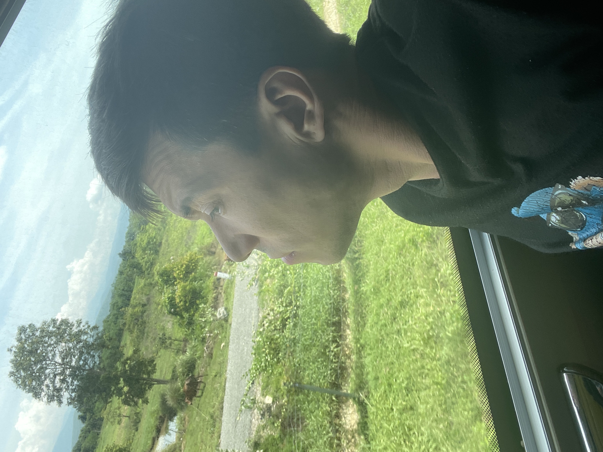 In Song Luy, Vietnam, a close-up profile shot captures a person looking out a window, with a rural landscape visible outside. The person, viewed from the right side of their face, has dark hair and is wearing a black hoodie with a graphic print. The window reveals a scene of rolling green hills, a single tree, a body of water, and a cow grazing in a field. A road runs through the middle ground. The sky is bright with scattered clouds. The interior of what appears to be a vehicle is visible at the bottom of the frame, including a portion of a door handle and the window frame.