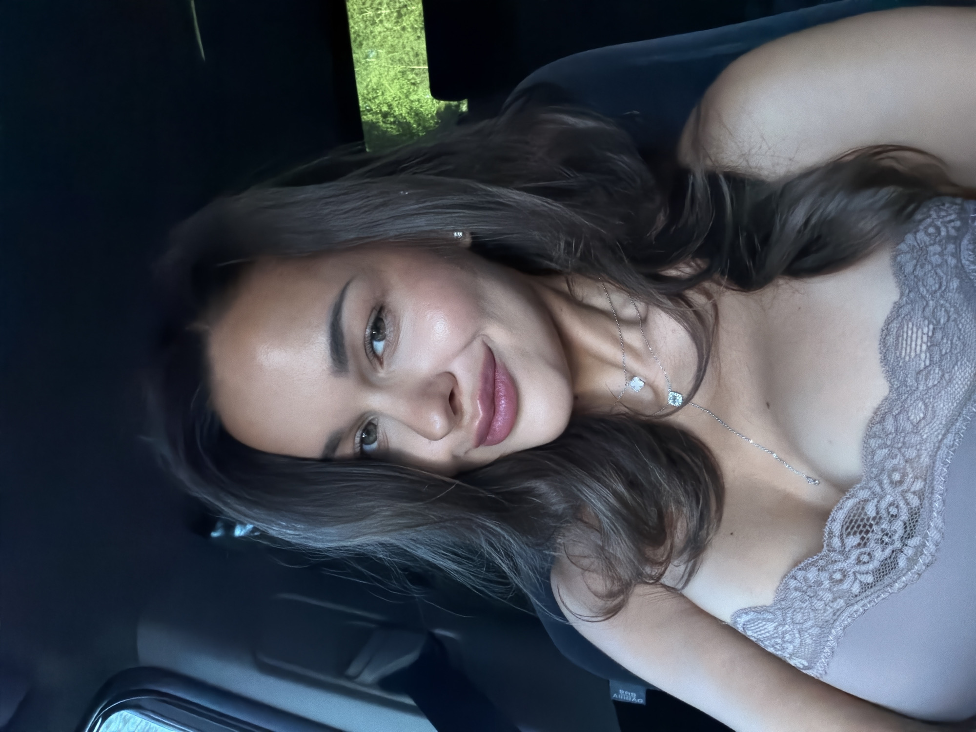 A young woman with brown hair, wearing a light-colored lace-trimmed top and jewelry, is lying in a vehicle. The background appears to show glimpses of greenery through a window. The location, Minino Stantsiya, Russia, is only a descriptor, and not visibly confirmed.