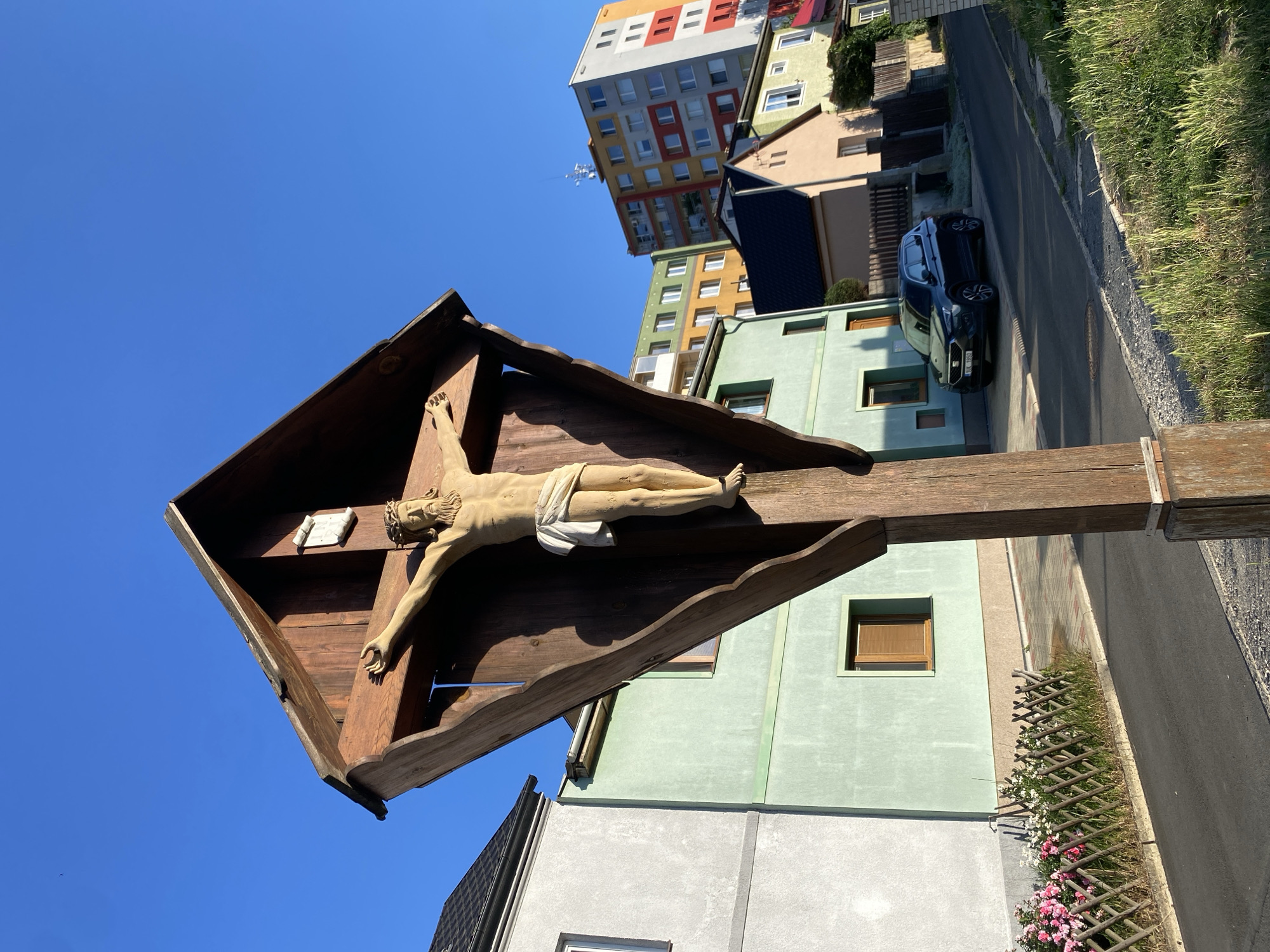 In Bílina, Czechia, a wooden crucifix with a statue of Jesus Christ stands prominently in front of a row of houses under a clear blue sky. The crucifix is mounted on a wooden post and features a triangular wooden roof. The statue of Jesus is light-skinned, wearing a white loincloth and a crown of thorns. Behind the crucifix, a two-story house painted light green is visible, with a single brown-framed window on its upper floor. To the left of the green house is a grey house with a dark roof. Further down the street, a dark blue SUV is parked on the asphalt road. Several other residential buildings, including a colorful apartment building with balconies, line the street. The scene captures a quiet residential area with a religious monument.