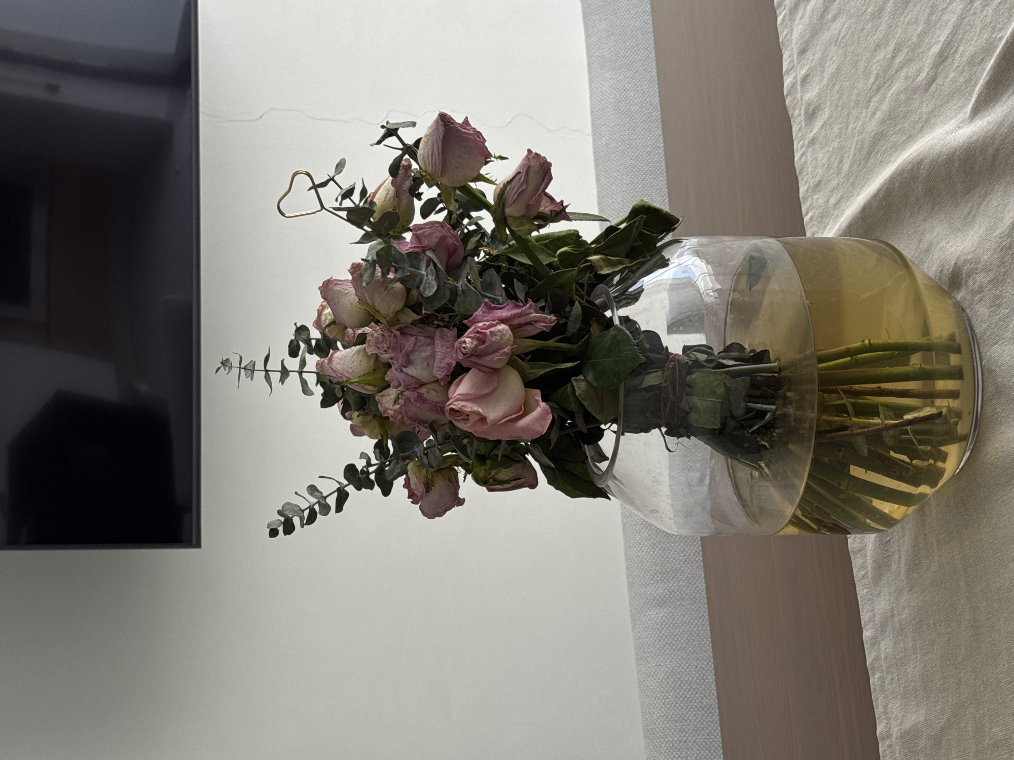 The image shows a bouquet of dried pink roses and greenery in a clear glass vase filled with yellowish water. The vase sits on a beige surface, possibly a bed or table, near a white wall and a black television. There is no information in the image to suggest that the location is Puebla, Mexico.