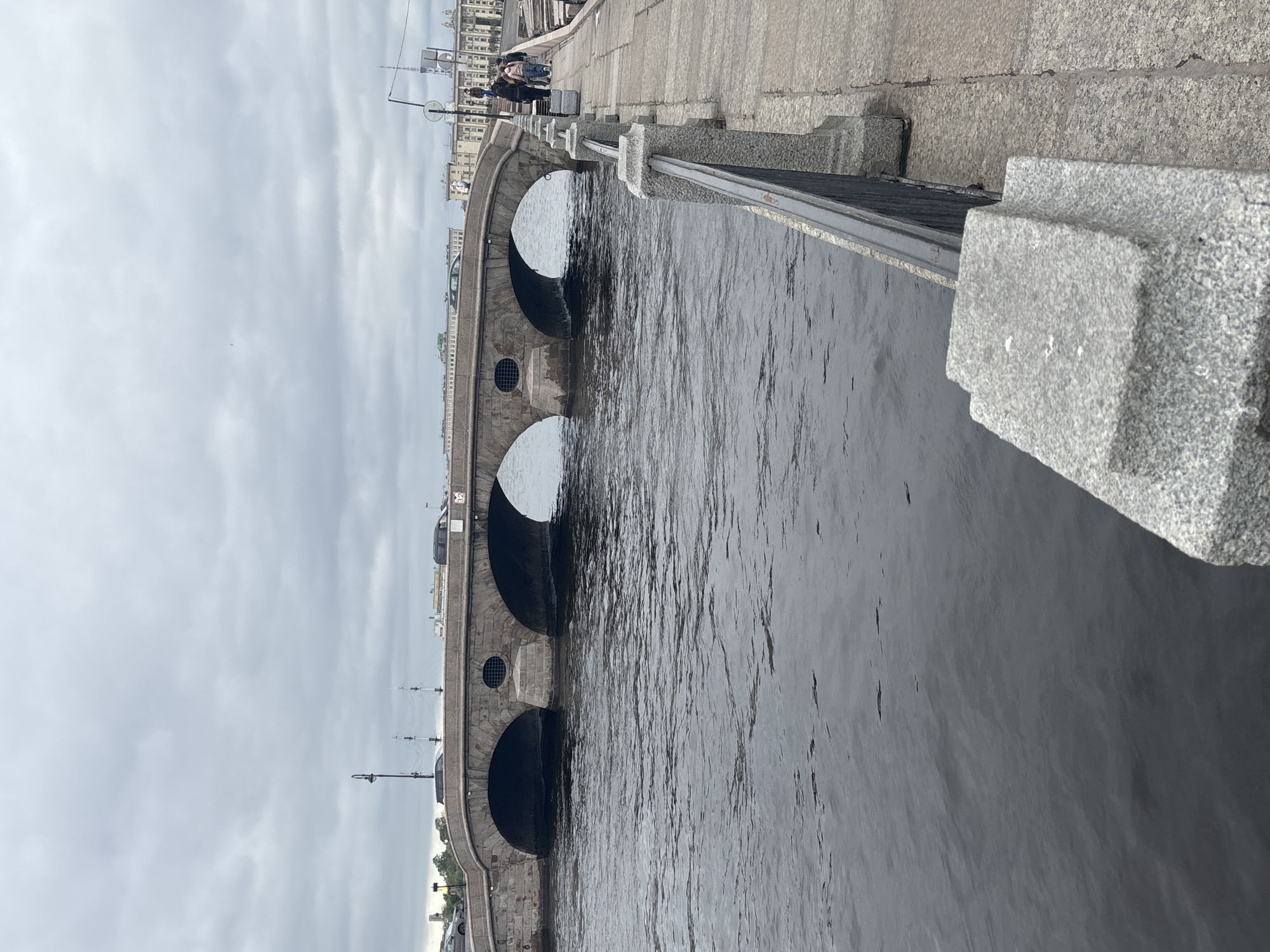 The image shows a stone bridge over a dark body of water in Saint Petersburg, Russia. The bridge has multiple arches and a granite balustrade along the top. People can be seen walking on the bridge. The sky is overcast. The water is dark and slightly rippled.