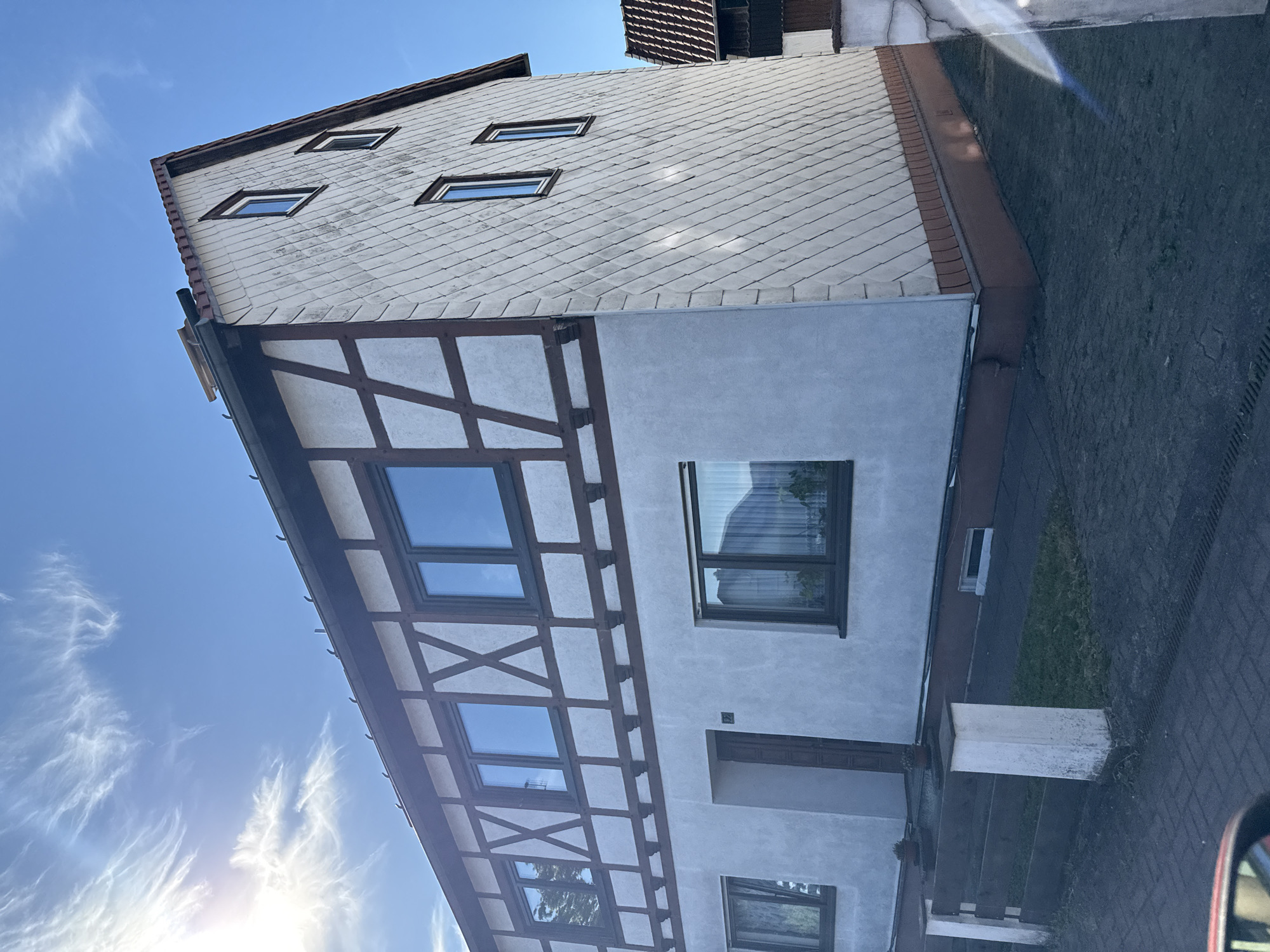 This image shows a two-story building in Bad Sachsa, Germany, under a clear blue sky with wispy clouds. The building features a distinct half-timbered facade on the upper level, with white infill between dark brown timbers arranged in geometric patterns. The lower level is plastered in a light grey color. The right side of the building is covered in light-colored diamond-shaped tiles, creating a textured appearance. Several windows are visible on both levels, with dark frames. A red-brown base runs along the bottom of the building, and a tiled roof is partially visible. In front of the building, a dark paved area is visible, bordered by a low wooden fence with white posts. Small potted plants are placed near the entrance. The overall scene depicts a residential building in a European town.