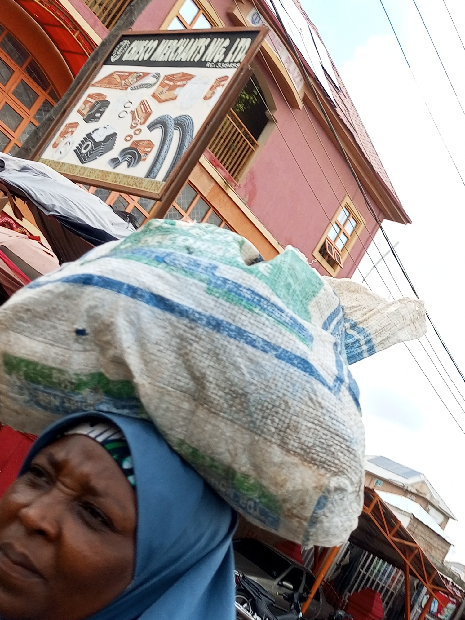 An upward-angled shot captures a woman wearing a blue hijab carrying a large, white sack with blue and green stripes on her head. She is positioned in the foreground, with her face in profile and her expression appearing strained. Behind her, a pink building with orange trim and wooden window frames is visible. A framed sign hangs on the building, displaying various automobile parts and the text "CRISCO MERCHANTS NIG. LTD". The sky is overcast. The scene is set in Arawa, Nigeria.