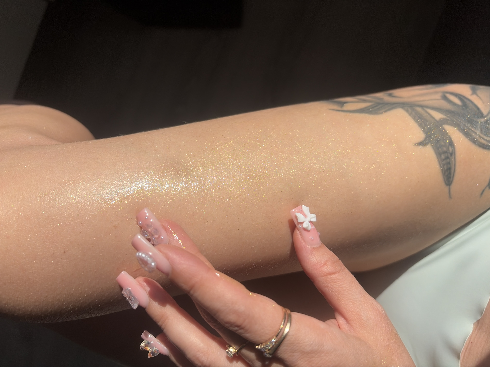 The image shows a close-up of a person's arm and hand. The skin is illuminated with a golden shimmer, suggesting the presence of body glitter or a shimmering lotion. The person has long, pink acrylic nails adorned with small pearls and translucent embellishments resembling butterflies and flowers. They are wearing two rings on one finger: a thinner gold band with small, prong-set clear stones and a thicker gold band with a raised, crown-like design. A tattoo is visible on the lower part of the arm, featuring dark, stylized designs that appear to be feathers or flames. The background is dark and out of focus, with a sliver of light illuminating the arm and hand. The location in Saint Petersburg, Russia, is not determinable from the visual information provided.