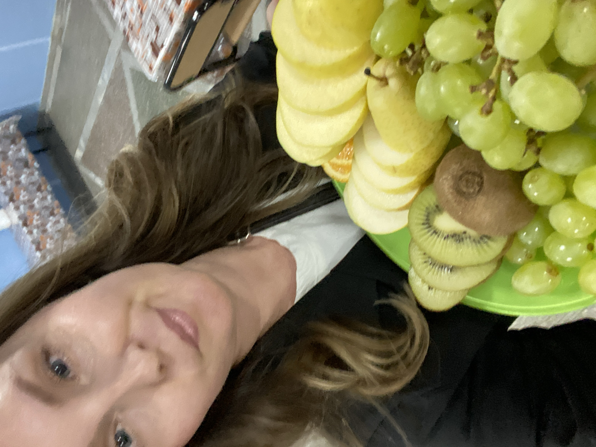 A person is holding a plate of sliced fruit, including kiwi, pears, and grapes. The person has long, wavy brown hair and blue eyes. The background includes what appears to be a tile floor and other items on a table. The city of Noviy, Russia, is not discernible from the image.