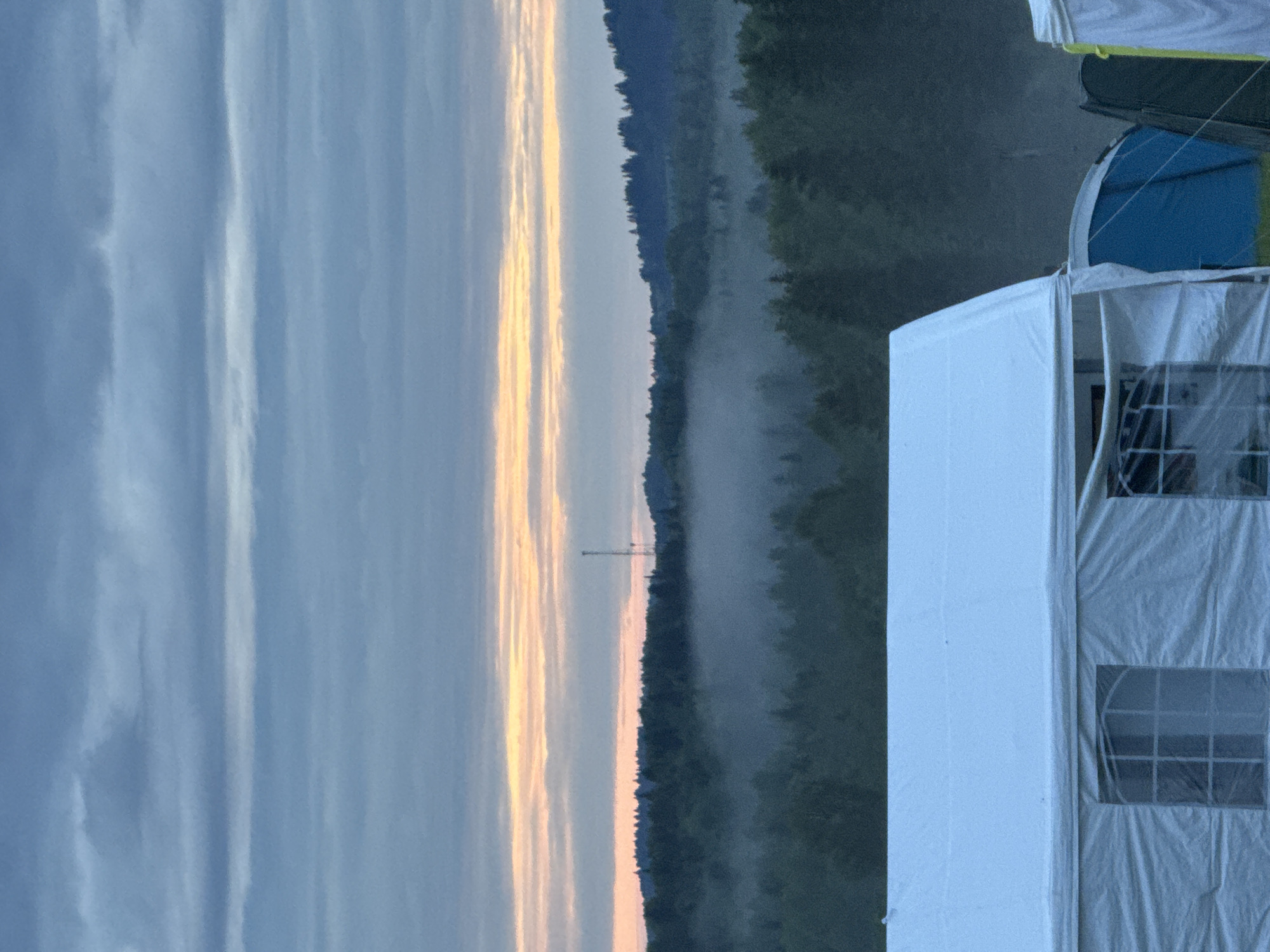 The scene is captured in Большая Кивара, Russia, at low light, suggestive of dawn or dusk. The sky, in the upper portion, displays stratified grey-blue clouds with a horizontal band of orange and pink light at its lower edge. A distant horizon of dense, dark green forest is visible, with a slender, dark tower silhouetted against the sky slightly right of center on the tree line. A mid-ground forested area is enveloped by a layer of ground fog. A large white tent or marquee structure occupies the lower half of the image, featuring window sections with visible grid patterns. To the right, parts of a blue dome tent and a dark-colored tent with yellow trim are partially visible.