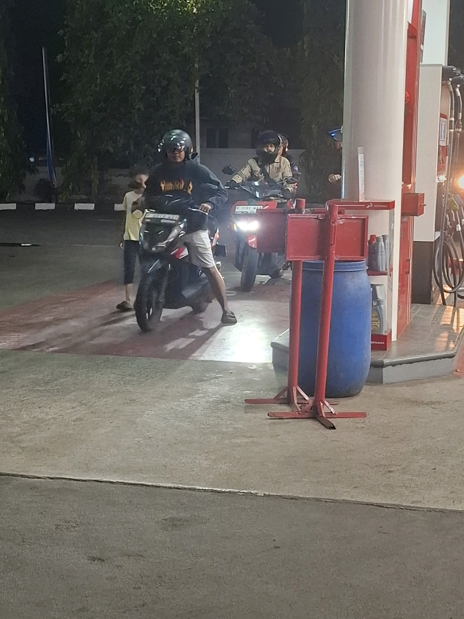 At a gas station in Karangtengah, Indonesia, two individuals on a scooter are parked near a fuel pump. The rider wears a dark hooded sweatshirt and a helmet, while the passenger, a child in a yellow shirt and dark pants, stands beside the scooter. Another person on a separate scooter, wearing a tan jacket and helmet, is parked behind them. A large blue barrel and red metal stands are visible near the fuel pump, with gas hoses hanging down. The ground is paved with concrete and marked with red lines. Trees and buildings are visible in the background. The scene appears to be at night, with artificial lighting illuminating the area.
