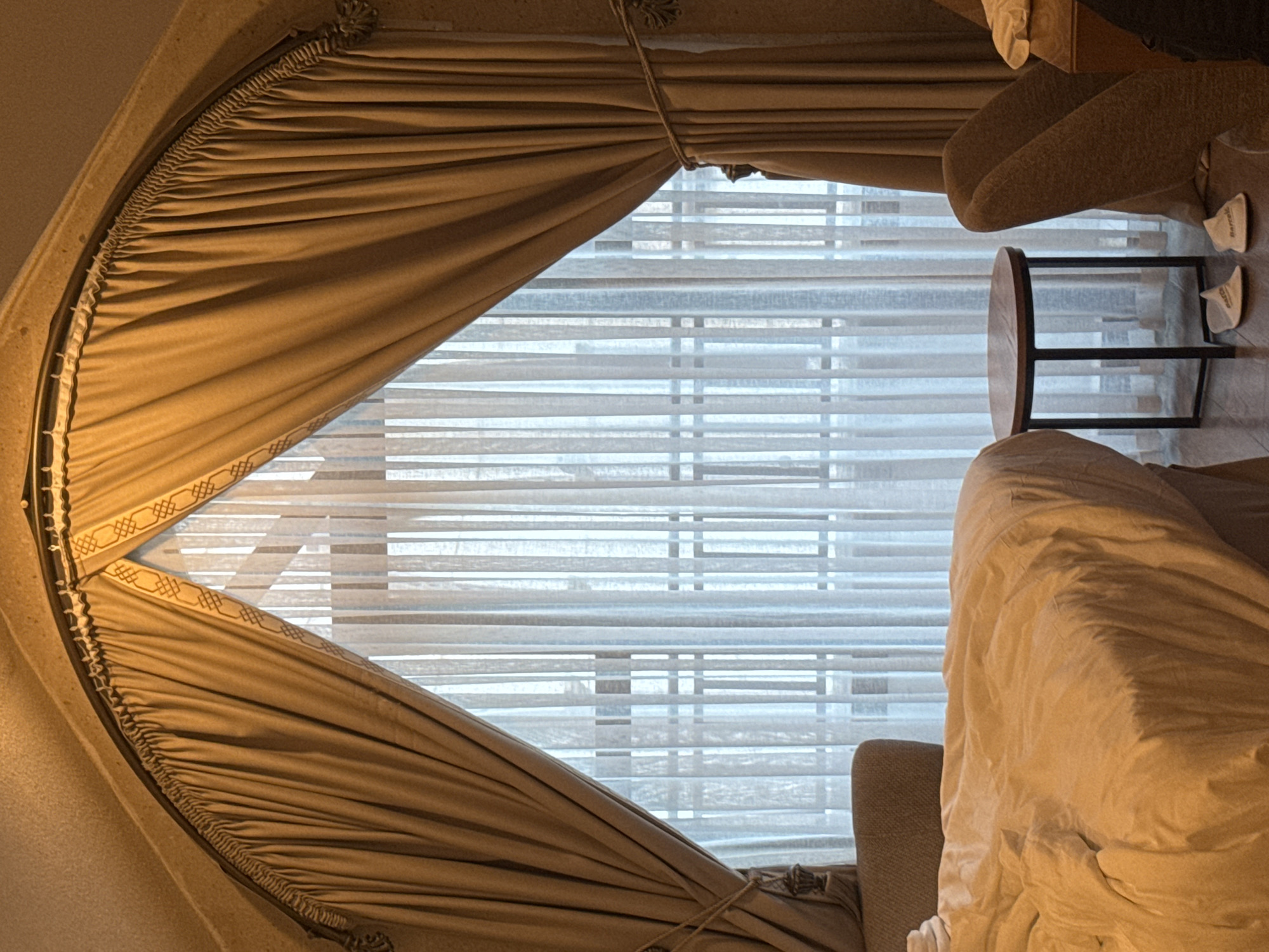 The image shows the interior of a room, possibly in a hotel or private residence. The focal point is a large window covered with sheer white horizontal blinds, partially obscured by ornate, heavy tan-colored drapes. The drapes are gathered at the top and sides, creating a diamond-shaped opening that reveals the blinds.
To the right of the window is a small round table with a dark metal frame and a light-colored wooden top. Next to the table are two pairs of white slippers. A portion of an upholstered chair is visible, partly covered with a rumpled white blanket or bedspread. The lighting in the room appears soft and diffused, creating a warm ambiance. The interior could be located in Ürgüp, Turkey, but there is nothing in the image that confirms it, it would be speculation.