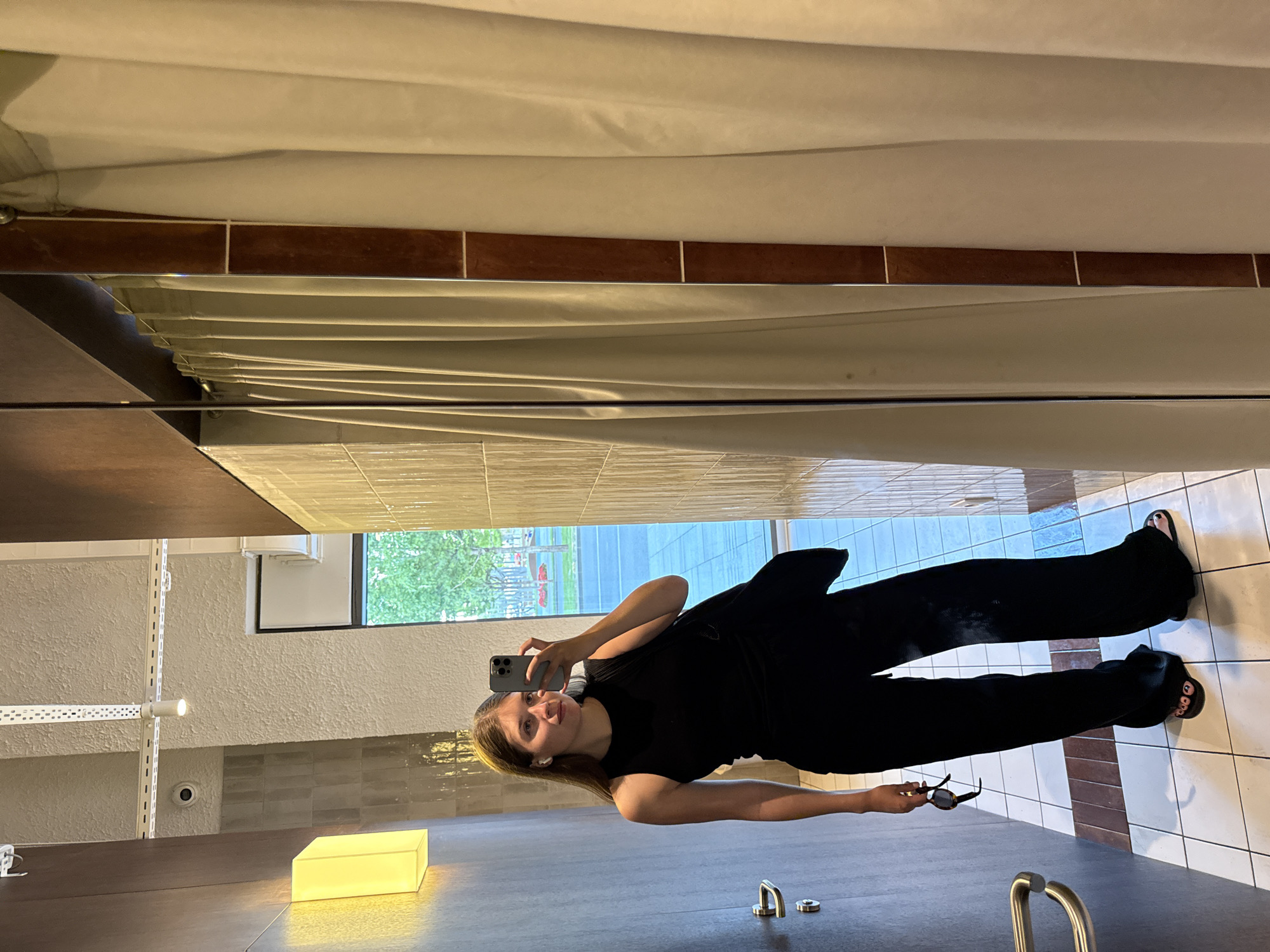 A woman is taking a selfie in a mirror. She is wearing a black outfit consisting of a top and wide-leg pants with open-toed sandals. She is holding sunglasses in her left hand and a smartphone in her right. The reflection in the mirror shows a room with light-colored tile walls, a window, and a drop ceiling with a curtain. The location is likely a retail store or dressing room in Belgrade, Serbia. There is a decorative light fixture, and chrome fixtures appear in the foreground.