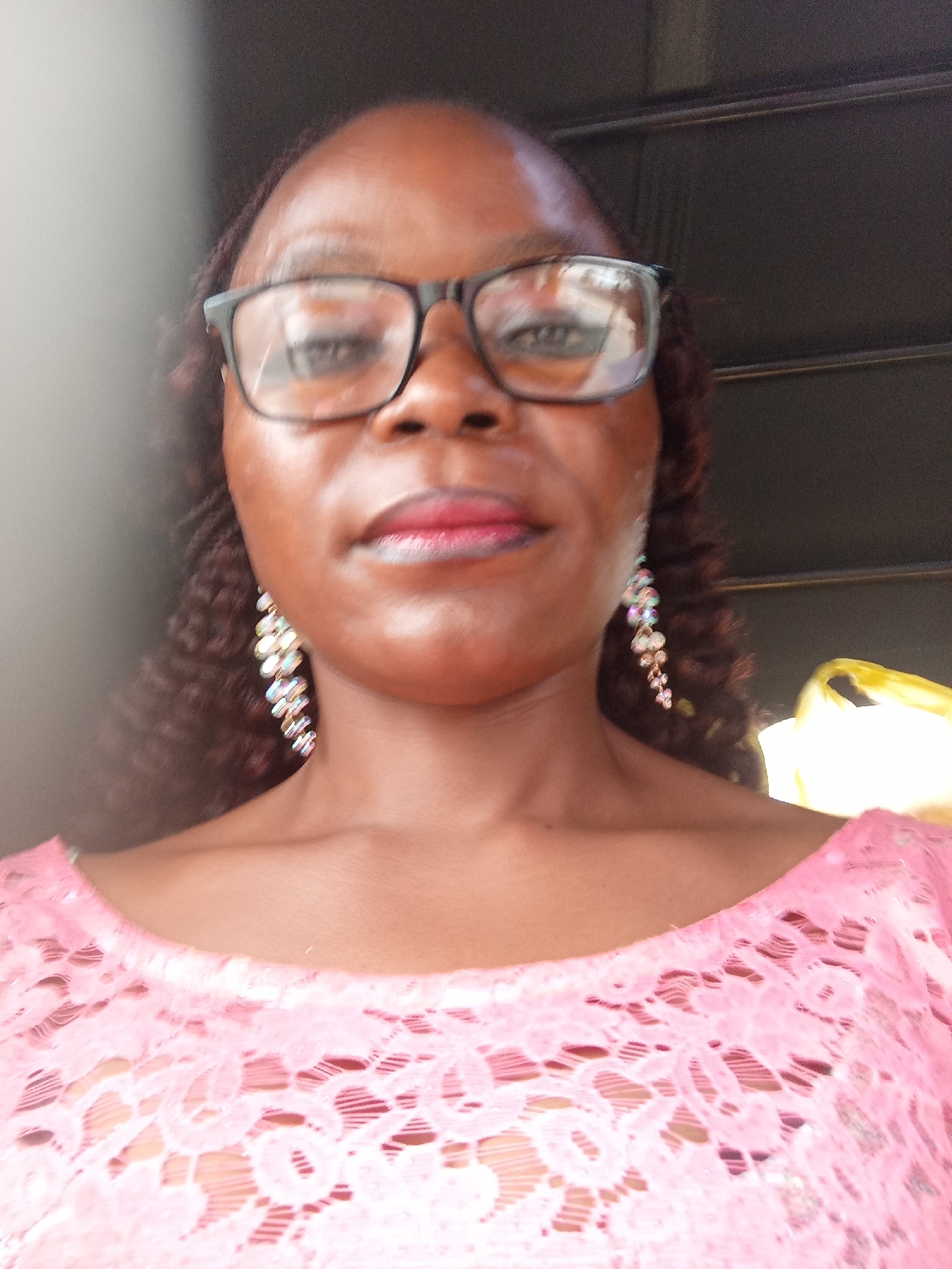 The image is a close-up portrait of a woman wearing glasses and a pink lace top. She has on large, dangling earrings. The background is out of focus but appears to be dark with some horizontal lines, possibly a textured wall or blinds. There is a bright yellow object to the right of the woman, partially visible. The woman's skin tone is dark. The lighting suggests she is outdoors or in a well-lit room. There are no visible landmarks or contextual clues to indicate the specific location within Jalingo, Nigeria.