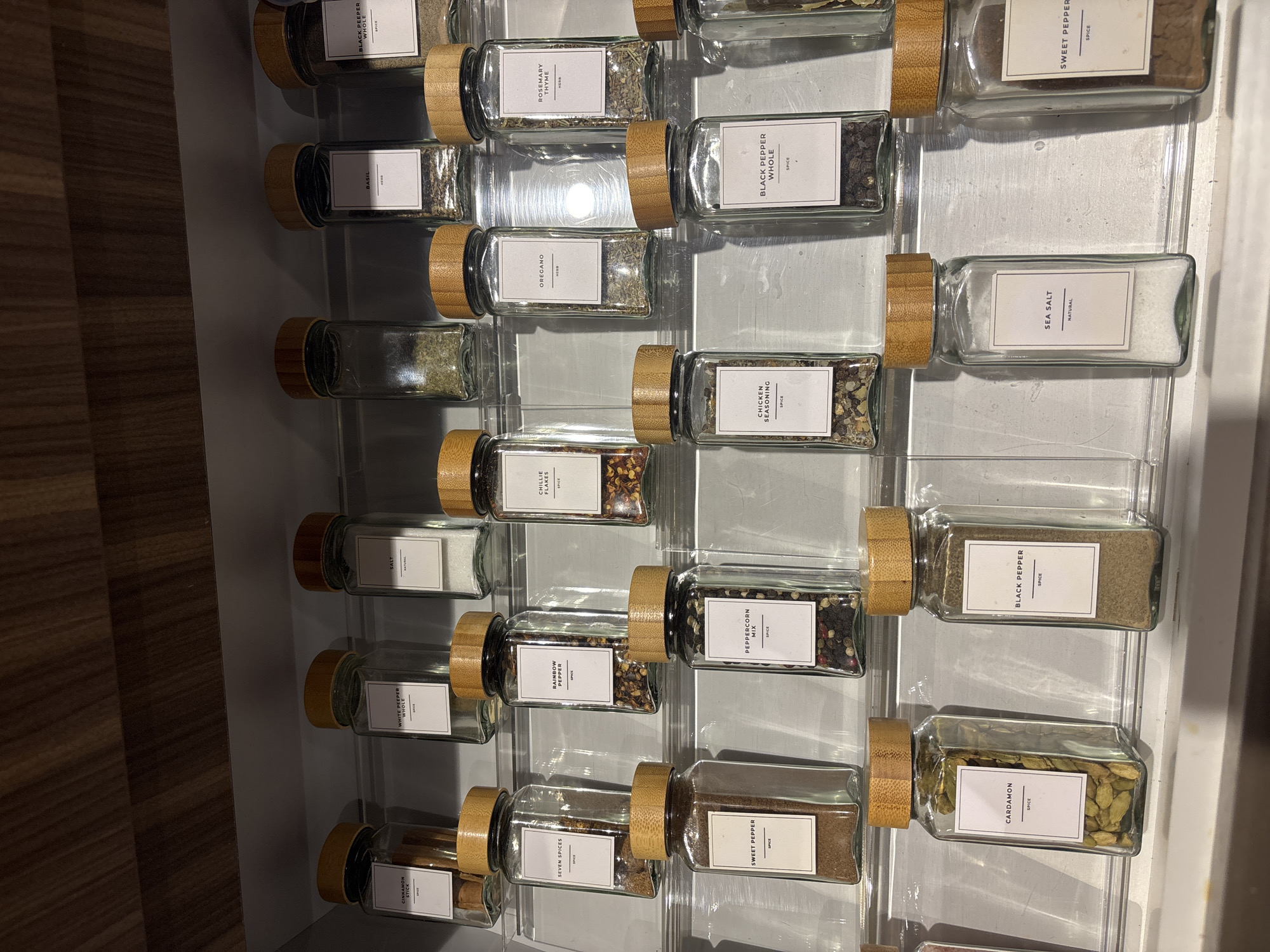 The image shows a close-up, eye-level view of a spice rack filled with numerous glass jars, each containing different spices. The jars have wooden lids and white labels with black text. They are organized neatly on multiple clear shelves. The environment appears to be an interior space, possibly a kitchen cabinet or pantry, with a wooden surface visible at the top. The lighting is even, illuminating the contents of the jars. The image does not contain any specific geographical markers or elements that would indicate a location in Dubai, United Arab Emirates. There are no people or actions depicted.