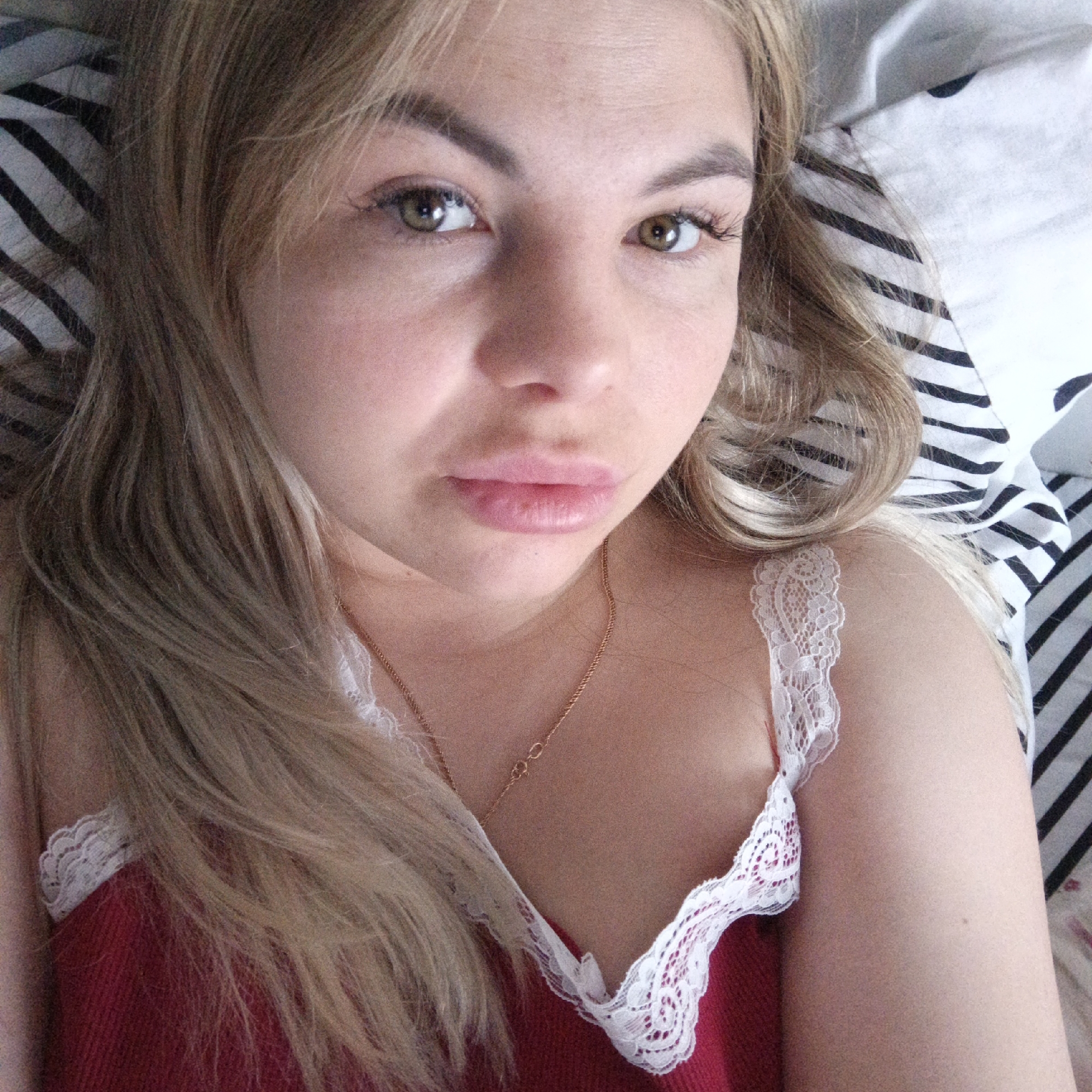 A close-up, high-angle shot depicts a woman lying down, looking directly at the camera. She has long, light blonde or light brown hair that cascades around her shoulders and frames her face. Her eyes appear to be a light brown or hazel color. Her eyebrows are well-defined, and she has visible eyelashes, suggesting mascara or extensions. Her lips are full and slightly pouted.
She is wearing a red camisole or top with delicate white lace trim along the neckline and straps. A thin, gold chain necklace is visible around her neck, with its clasp positioned near her collarbone.
The woman is positioned on what appears to be a bed, partially covered by bedding with distinct black and white stripes. The lighting suggests an indoor setting, likely natural light from a window. The overall mood is casual and intimate, possibly a self-portrait taken in a relaxed moment. There is no visible text in the image.