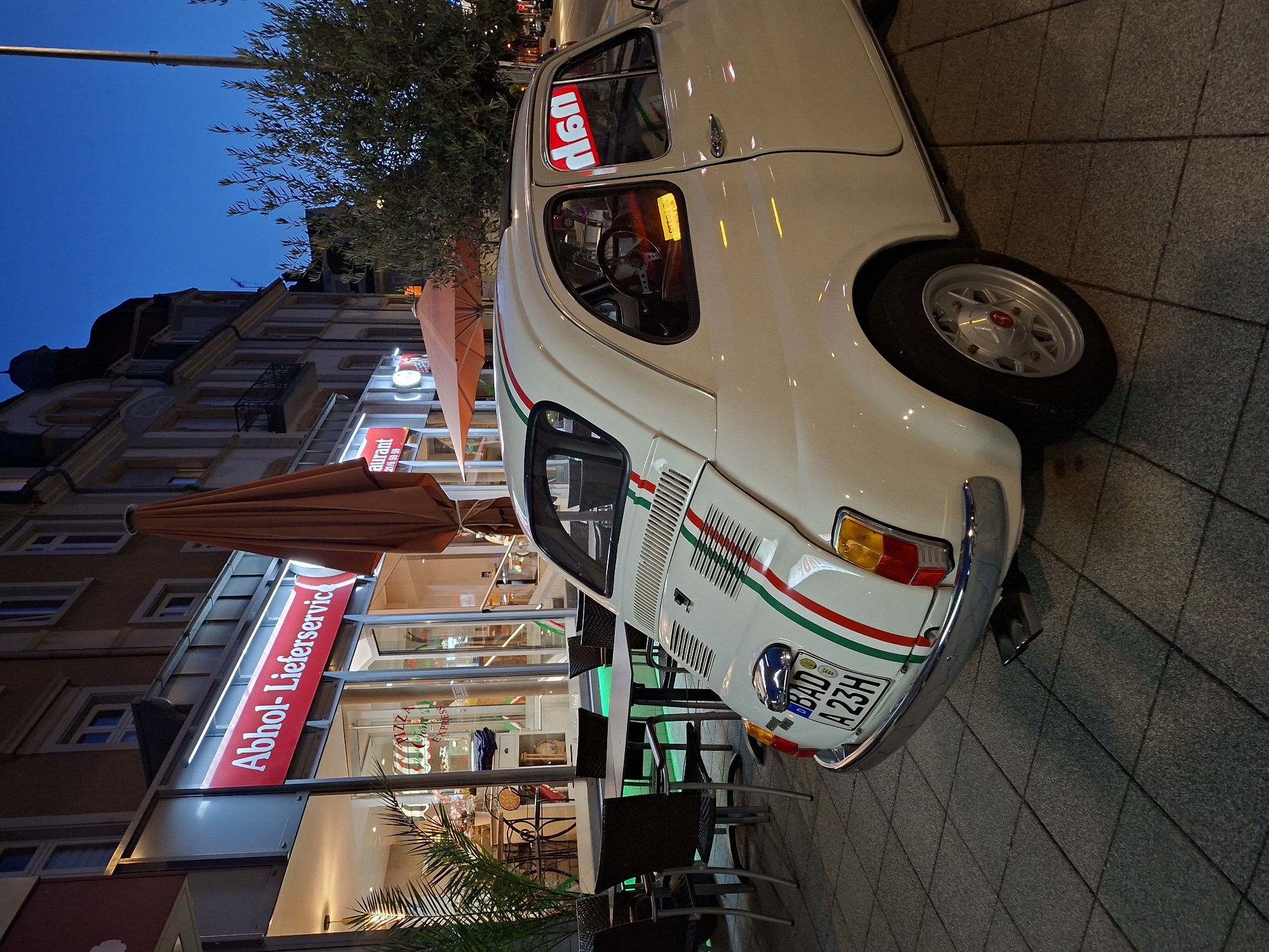 A white Fiat 500 car, with license plate "BAD A23H", is parked on a tiled sidewalk in front of a restaurant at dusk in Baden-Baden, Germany. The restaurant, identified by a red sign reading "Abhol-Lieferservice" (Takeaway-Delivery Service), has large glass windows and outdoor seating with tables and chairs. An orange umbrella and a large brown umbrella are also visible outside the restaurant. The car is decorated with red, white, and green stripes on its rear, and a red "06n" sticker is visible in the passenger side window. The street is lined with buildings, and the sky is a deep blue, indicating twilight.