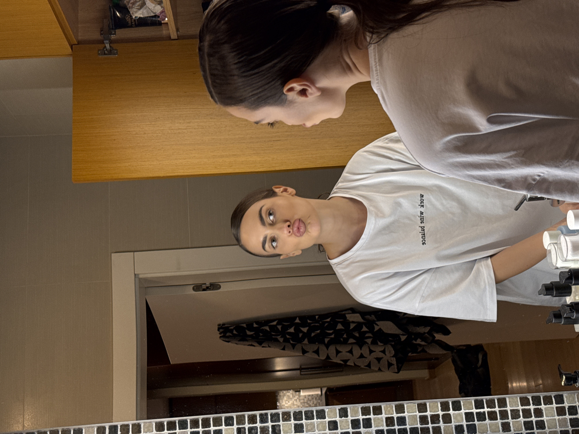 A photograph captures a woman reflected in a bathroom mirror. She is facing the mirror with her lips puckered in a kissing gesture, wearing a white t-shirt with black lettering. Behind her, a glimpse of a light wood cabinet is visible along with toiletries. The mirror frame is decorated with small mosaic tiles. The location is not identified within the image.