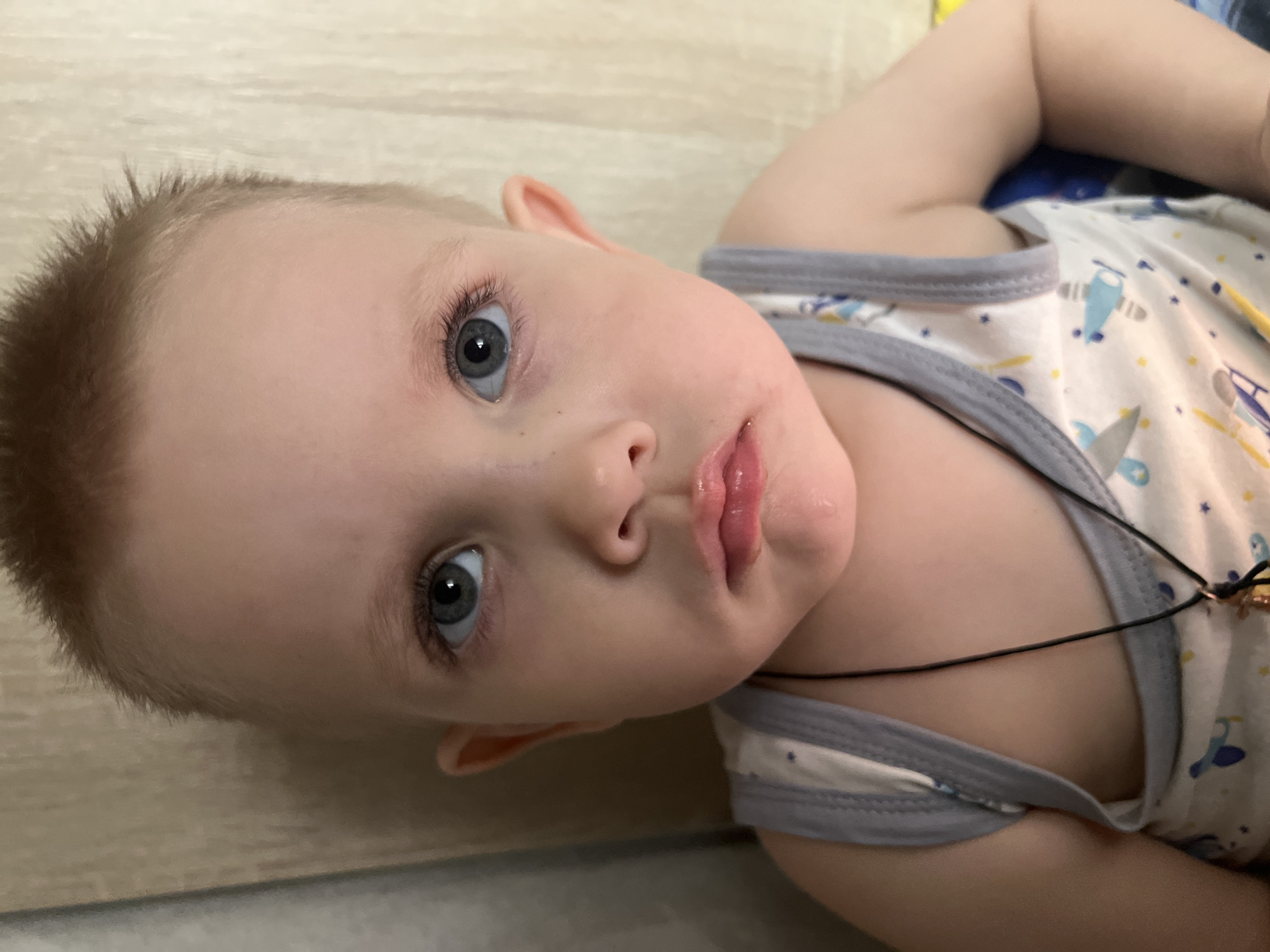 A close-up shot depicts a young child with light hair and blue eyes lying on a surface. The child is wearing a white and grey trimmed tank top with an airplane pattern, and a thin black cord is visible around the child's neck. The location is unspecified and the city, Марковский, Russia, is not evident from the image.