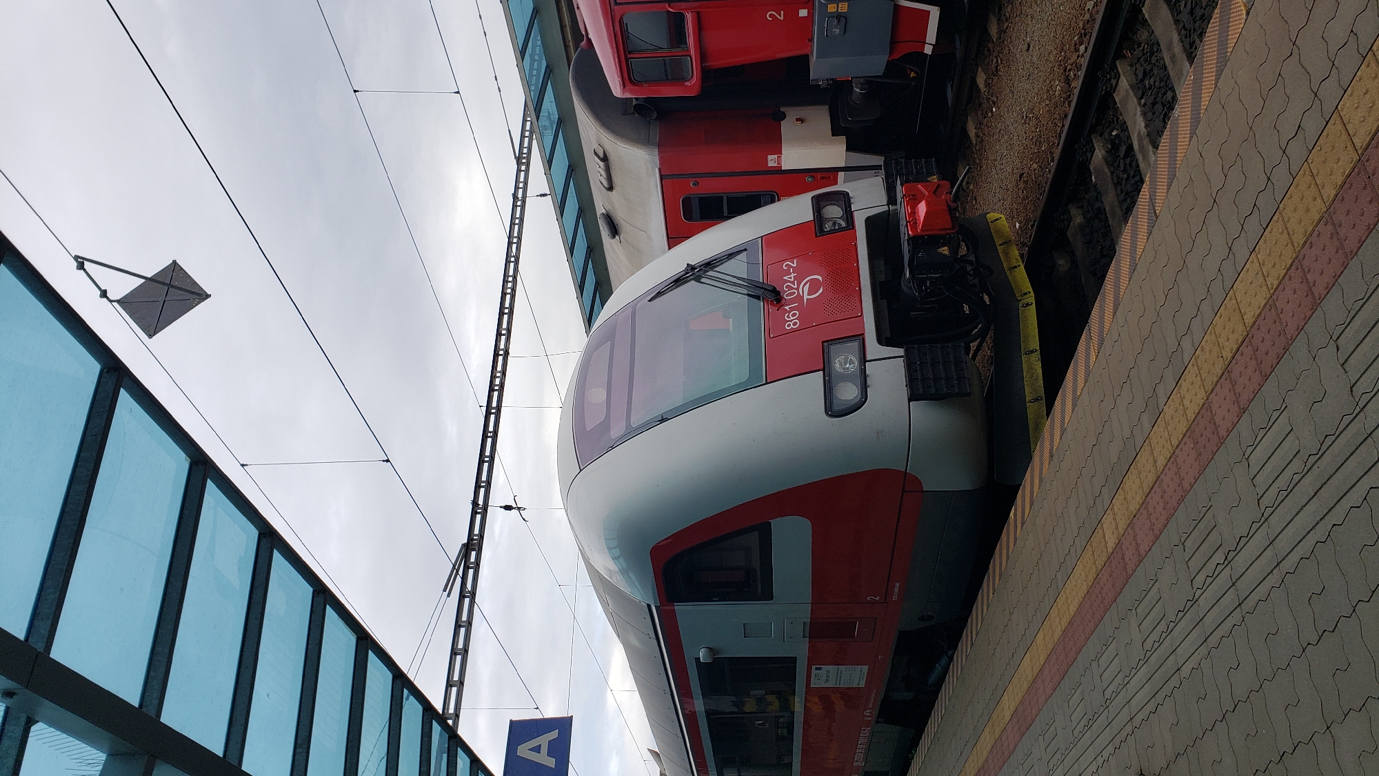 The image depicts a train at a station platform. The train is predominantly red and white, with a visible number "861 024-2" on its front. It is parked on a railway track, adjacent to a platform with textured paving stones in yellow, red, and grey. Overhead, a blue glass canopy supported by metal beams covers the platform. The sky is overcast. In the background, another red train car is visible. The scene is located at the Prešov railway station in Prešov, Slovakia. There are no people visible in the image.