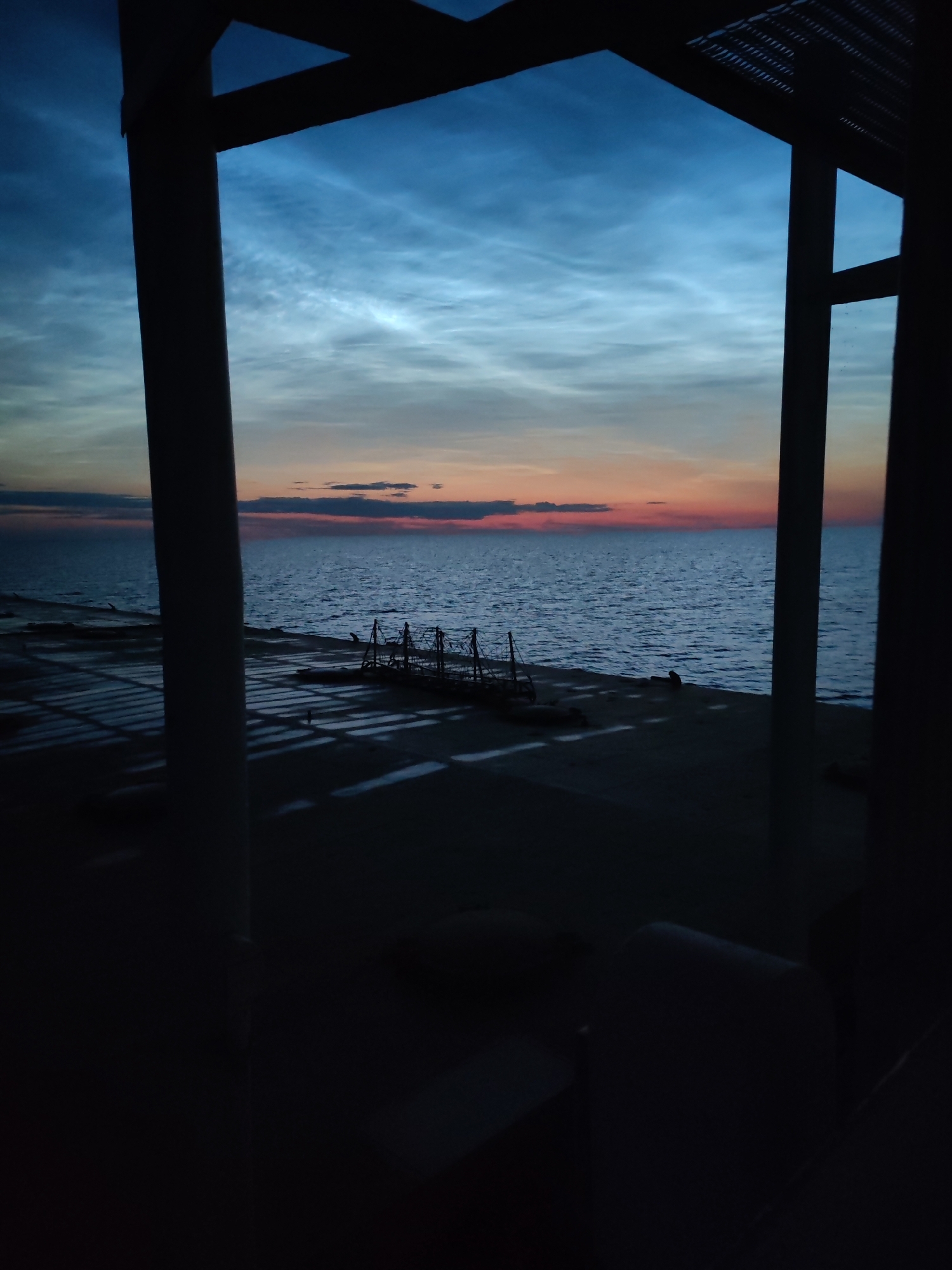 The image depicts a twilight scene viewed through a dark, silhouetted structure, likely an open-air pavilion or overhang, in Ozernoe Ust'e, Russia. The sky is a gradient of deep blue at the top, transitioning to lighter blues and streaks of white clouds, with a band of soft orange and pink near the horizon. Below the sky is a body of water, appearing to be a large lake or sea, with gentle ripples reflecting the colors of the sky.
The foreground is dark, with the prominent vertical and horizontal elements of the viewing structure obscuring the lower part of the frame. On the paved area between the structure and the water, there are several silhouetted objects. To the left, there are geometric shadows cast on the ground. In the middle ground, a cluster of dark, angular structures, possibly scaffolding or equipment, are visible. Further to the right, a single, dark human figure is seated or crouched on the ground. The overall impression is one of a serene, calm evening setting by the water.