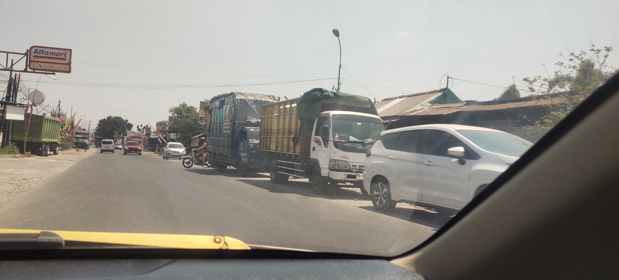 The image depicts a street scene in Cabean, Indonesia, under a bright, hazy sky. The perspective is from inside a vehicle, looking forward. On the left side of the road, a green dump truck is parked, and further down, a white van is visible. The road itself is lined with various vehicles, including several trucks. One blue truck with a tarp-covered load is prominent in the middle ground, alongside a white truck with a yellow tarp. A white car is seen in the foreground, partially obscuring the view of the road and other vehicles. Several motorcycles and smaller cars are also present, contributing to a sense of traffic. In the background, buildings with corrugated metal roofs and some trees can be seen. A red sign for "Alfamart" is visible on the left, indicating a commercial presence. The overall impression is one of a busy, possibly congested, local road.