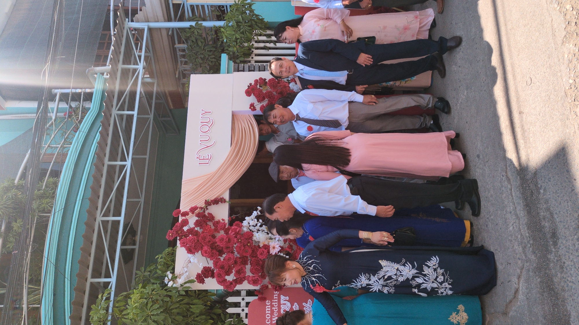 The image shows a group of people standing in front of a wedding arch in Phan Rang – Tháp Chàm, Vietnam. The arch is decorated with red and white flowers and a sign that reads "Lễ Vu Quy." The people are dressed in a variety of formal attire, including suits, dresses, and traditional Vietnamese áo dài. The environment is bright and sunny. The group appears to be posing for a photo.