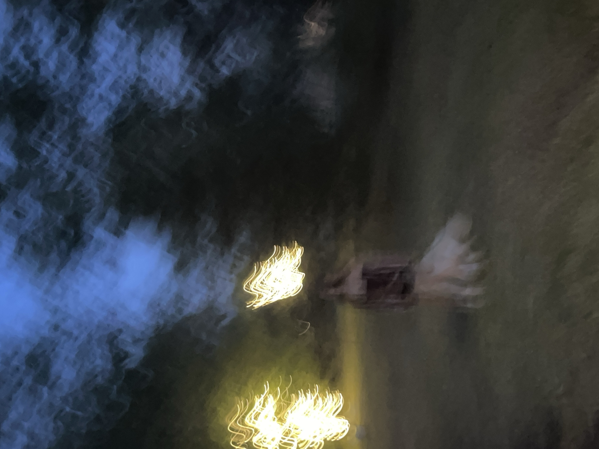 This is a blurry, abstract image taken at night, likely outdoors in Krasnoyarsk, Russia. The dominant colors are dark blues and greens, with streaks of bright yellow light. The yellow streaks appear to be from moving lights, possibly fireworks or light trails from vehicles, given their wavy, elongated shapes. There is a suggestion of a structure or object in the mid-ground, possibly a building or an installation, with some lighter, feathery shapes extending from it, which could be part of the object or another element in motion. The overall impression is one of movement and light against a dark, possibly natural, background. The time of day is clearly night due to the darkness and the artificial lights. There are no discernible people, text, or clear indications of weather, although the soft, diffused quality of the light and colors might suggest a slightly hazy or misty atmosphere. The abstract nature of the image makes it difficult to pinpoint specific objects or activities with certainty, but the dynamic streaks of light are the most prominent feature.