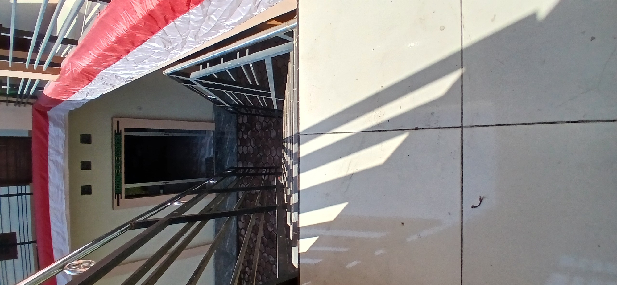 The image captures an outdoor view of a building entrance area in Pejaten, Indonesia, with a prominent staircase leading down. The building's facade is a light yellow, adorned with a red and white banner draped along the top edge, likely indicating a celebration or national holiday. Three square vents are visible above a doorway, which is partially open, revealing a dark interior. The staircase itself is constructed with metal railings on both sides and has tiled steps, some of which are illuminated by strong sunlight casting distinct shadows. The ground at the bottom of the stairs is paved with hexagonal tiles. The foreground shows a tiled surface with two dark, parallel grout lines, also partially covered by the shadows from the staircase. A small, dark, coiled object, possibly a piece of debris or a small insect, is visible on the foreground tiles. The overall scene suggests a sunny day with good lighting conditions.