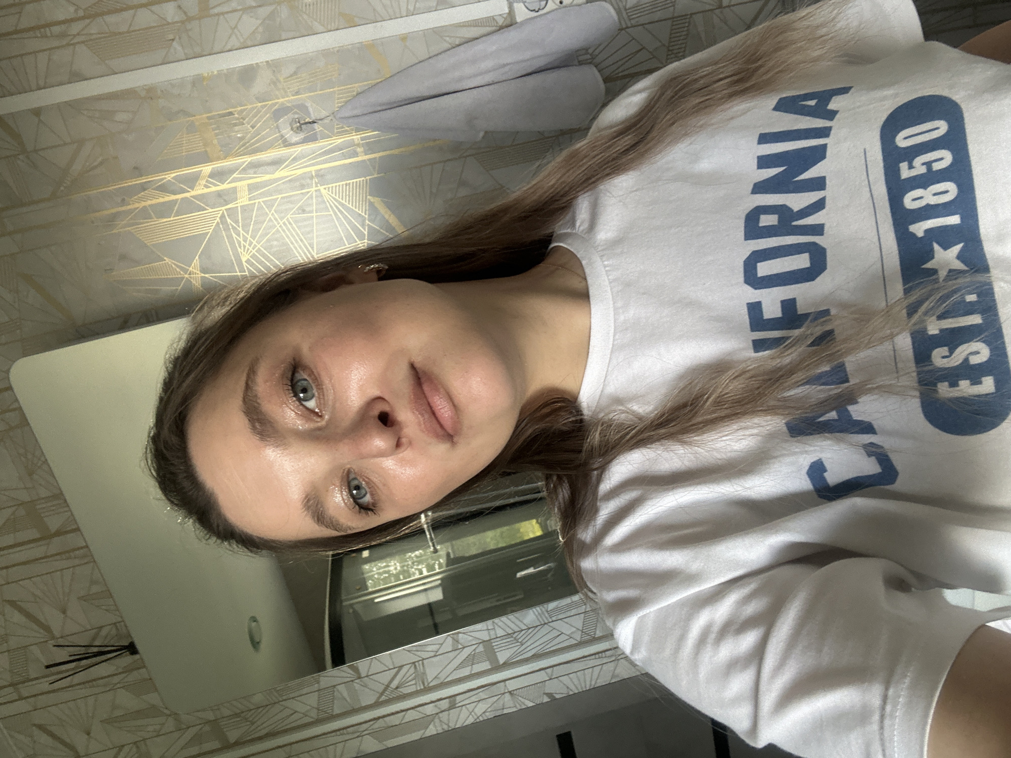The image shows a young Caucasian woman with long blonde hair, wearing a white t-shirt with blue lettering that reads "CALIFORNIA EST. 1850." She has blue eyes and fair skin with a slight shimmer, suggesting makeup or natural radiance. The woman is looking directly at the camera with a neutral expression. She is positioned in what appears to be a bathroom, indicated by the tiled walls with a gold geometric pattern, a mirror reflecting a doorway and possibly a shower or closet, and a towel hanging on the wall. The lighting is natural, coming from the left and reflecting off her face and the mirror. No specific landmarks of Вознесенка, Russia, are visible, and the scene is confined to the interior of what appears to be a residential or hotel bathroom.