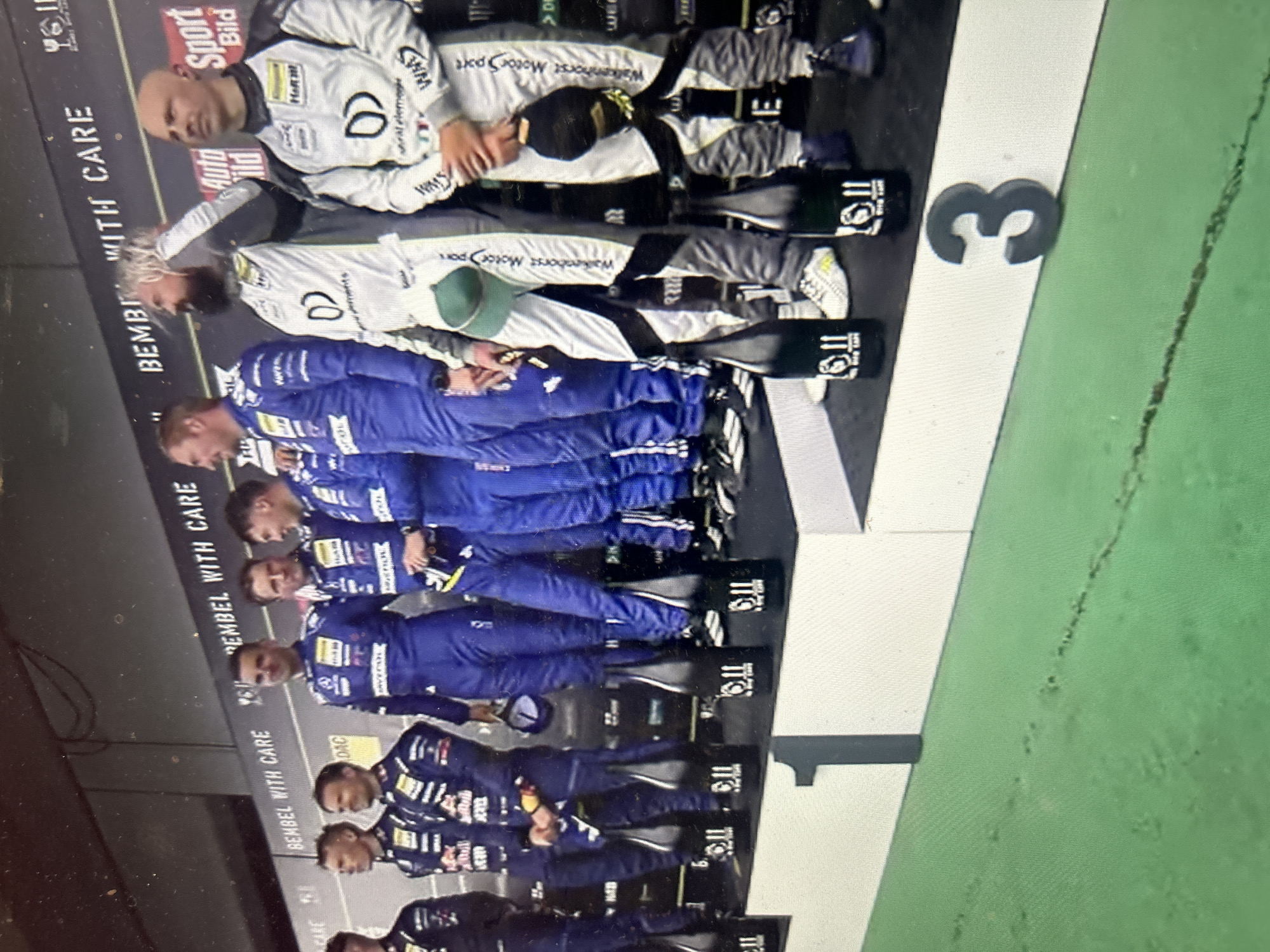The image depicts a group of individuals in racing suits standing on a podium with "1", "2", and "3" marked on the different levels. The first place is occupied by a group of people in blue suits. The second and third places are occupied by people in white racing suits with "Walkenhorst Motor Sport" printed on them. The brand "BEMBEL WITH CARE" can be seen in the background. The image does not show any location information of Sofia, Bulgaria.