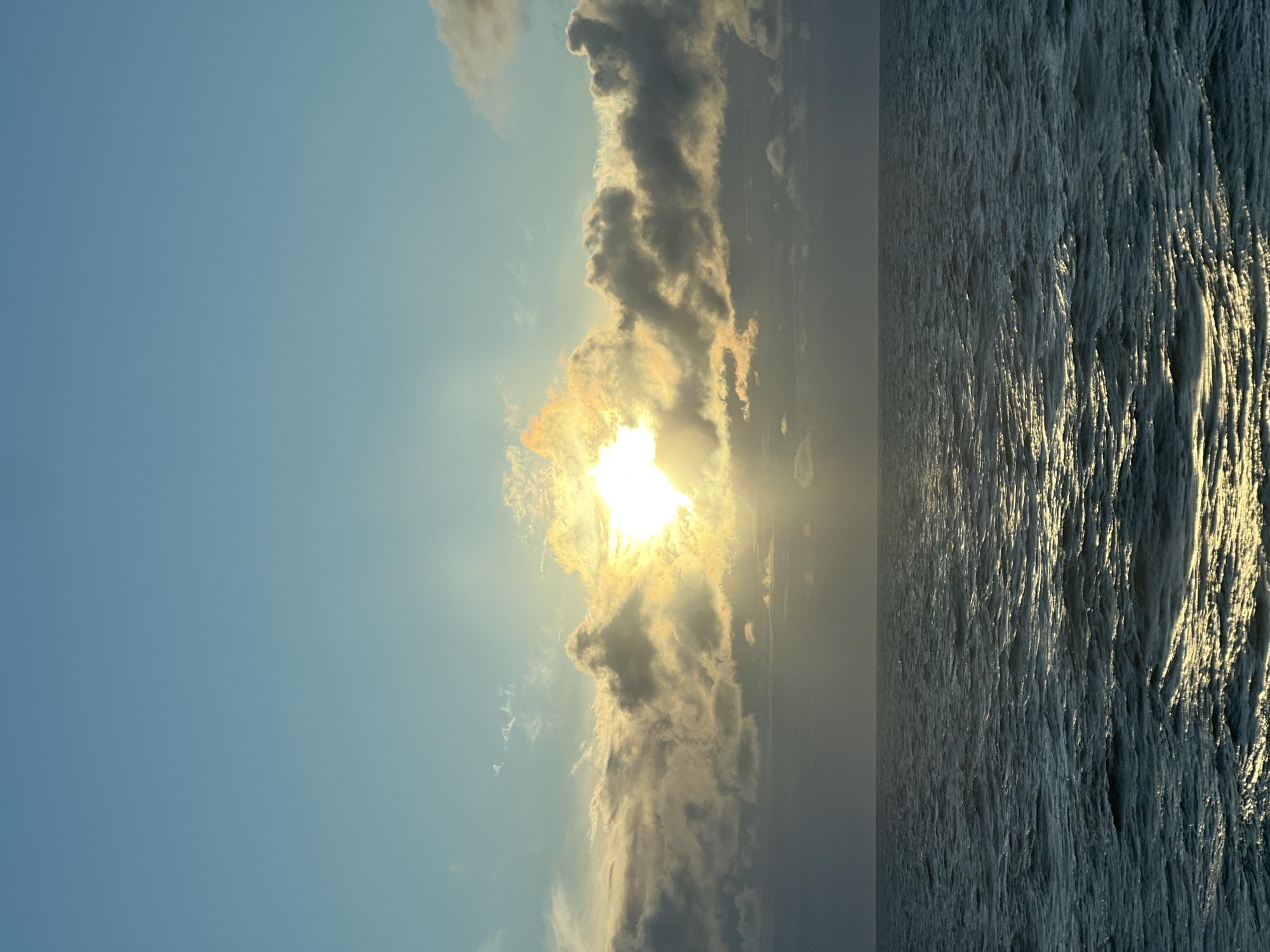 The image depicts a serene seascape at dawn or dusk. The sun, partially obscured by clouds, casts a warm, golden glow across the sky and reflects brilliantly on the rippling surface of the ocean. The sky transitions from a clear, pale blue at the top to a soft haze near the horizon, where the sun's rays break through. Darker, more textured clouds are clustered around the sun, creating a dramatic contrast. The ocean itself is a deep, undulating blue-green, with numerous small waves creating patterns of light and shadow on its surface. The horizon line is clear and sharp, dividing the vast expanse of the sky from the equally vast expanse of the sea. There are no visible human figures, vessels, or landforms in the image, making it a purely natural scene.