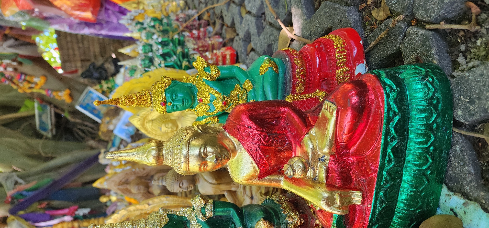 In Baan Chaweng Noi, Thailand, a close-up, eye-level view captures a collection of small, ornate Buddha statues arranged on a textured, grey stone surface. The foreground is dominated by two prominent statues. The leftmost is a gold-colored Buddha adorned in a shimmering red robe, seated with its hands resting on its lap, holding a small golden pouch. This statue is positioned on a layered base, the lower portion of which is a vibrant, metallic green with embossed floral patterns. To its right stands a green Buddha, also seated, wearing a golden headdress and elaborate golden jewelry around its neck and shoulders. This statue rests on a red base adorned with intricate gold detailing. Both statues have a slightly aged appearance, with some chips and wear visible on their bases. In the background, numerous other Buddha statues, varying in color and size, are visible, some appearing golden and others in shades of green. Behind the statues, the trunk of a large tree is partially obscured by colorful fabric streamers and garlands, including purple, pink, yellow, and green. A few small, framed images or cards are also attached to the tree trunk. The ground is scattered with dry leaves and twigs, suggesting an outdoor setting, likely a shrine or place of worship. The lighting is bright, casting subtle shadows.