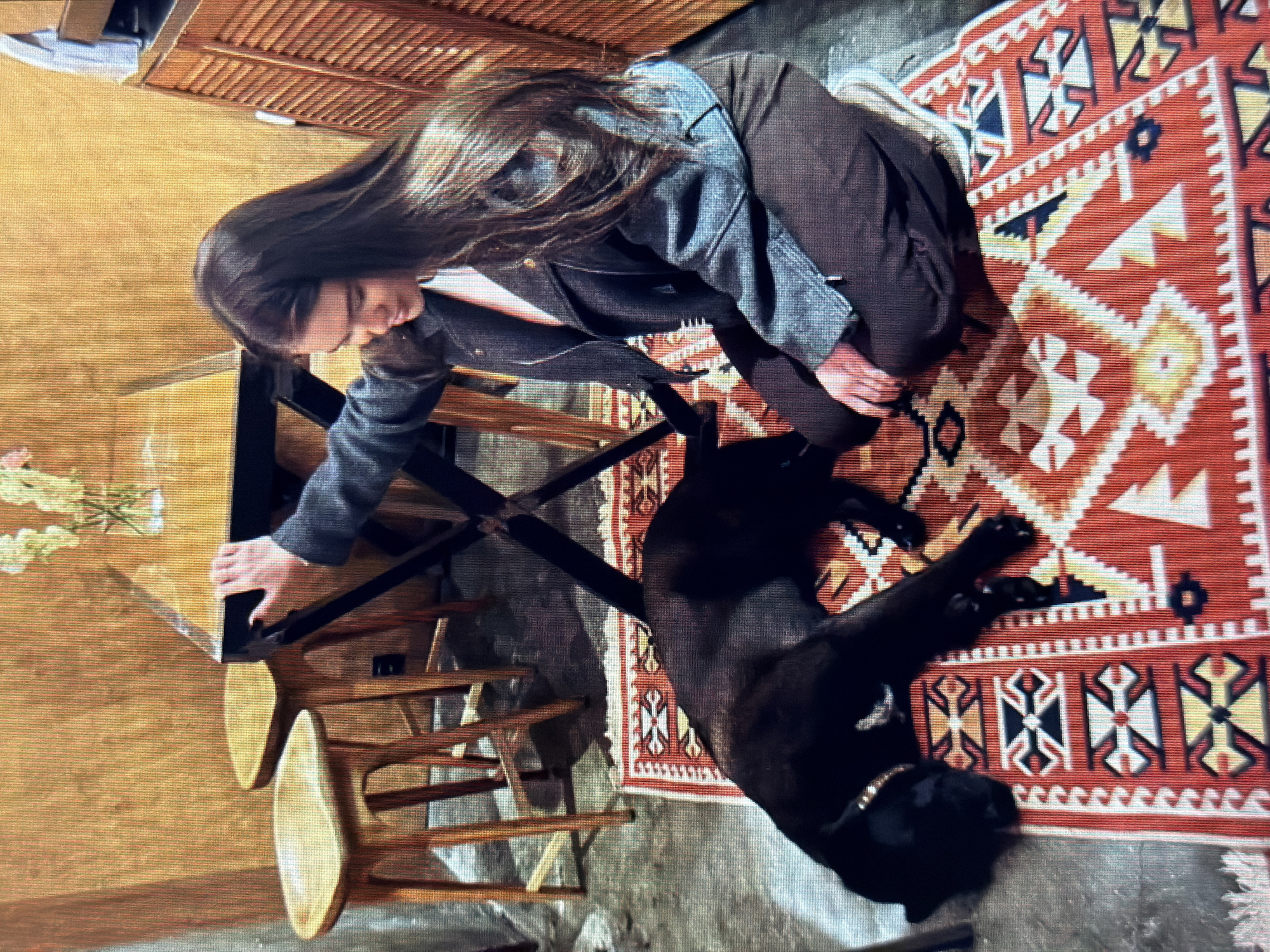 In an indoor setting, a young woman with long, dark hair is kneeling on a rug. She is wearing a grey jacket and brown pants. Her right hand is resting on a table. A black dog is lying down on the rug in front of her. The environment appears to be a cafe or restaurant, with wooden chairs and a table visible in the background. The wall behind the subjects is made of wood. The flooring is a mixture of concrete and the patterned rug. The image does not provide any information to determine the location within Sirius, Russia.