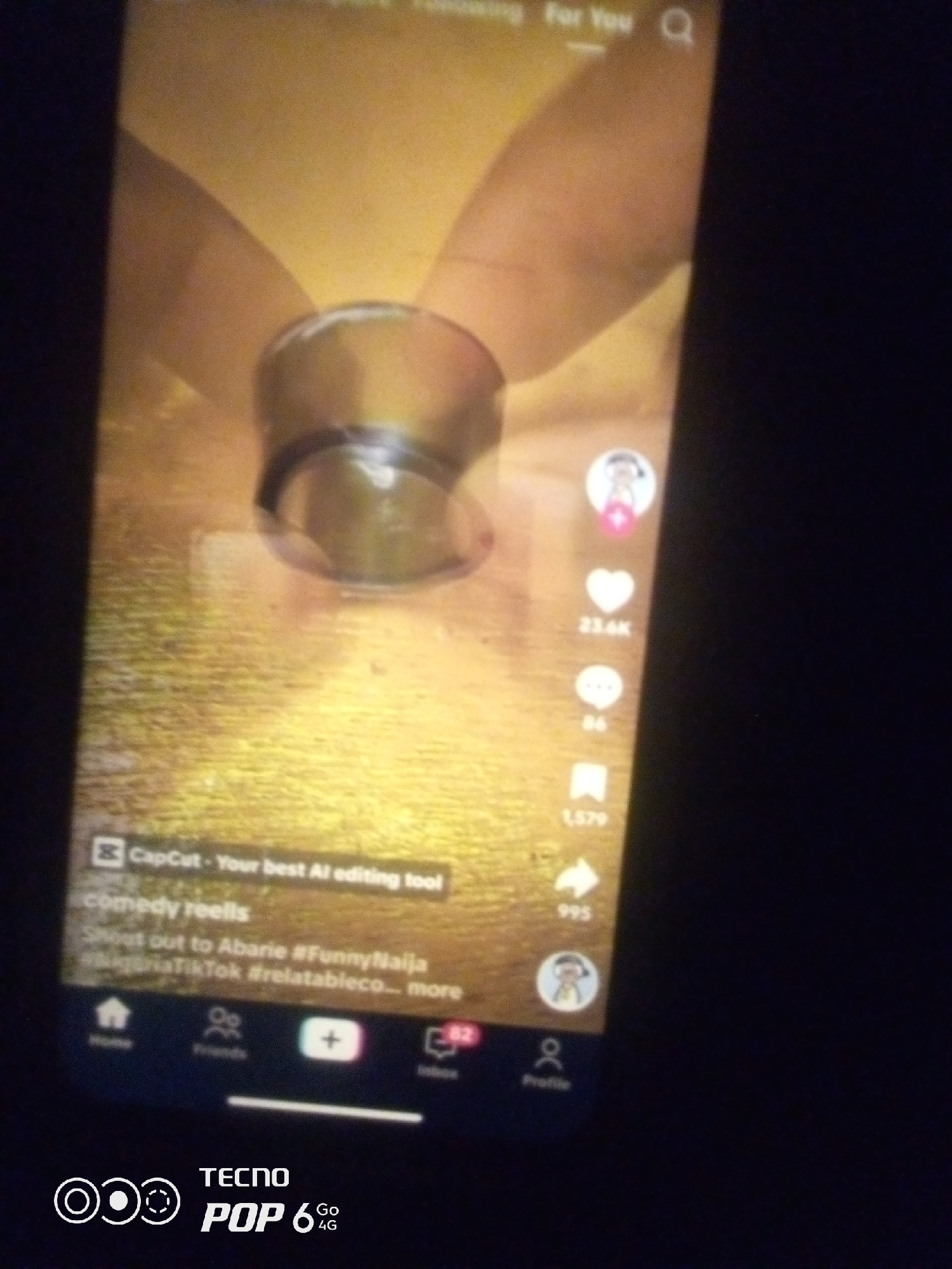 The image shows a close-up of a smartphone screen displaying a TikTok video. The video appears to be a comedy reel, with text overlays indicating "CapCut - Your best AI editing tool" and "comedy reels". There are also hashtags visible: "#Abarie", "#FunnyNaija", "#NigeriaTikTok", and "#relatableco...". On the right side of the screen, there are typical TikTok engagement icons: a heart with "23.6K" likes, a speech bubble with "86" comments, a bookmark icon with "1,579" saves, and a share icon with "995" shares. The bottom of the screen shows navigation icons for "Home", "Friends", a plus button, "Inbox" with "82", and "Profile". A Tecno Pop 6 4G logo is visible in the bottom left corner of the screen. The background of the video itself is a textured, yellowish surface with a blurred out object in the center, possibly a fan or a spinning toy. The image is taken in low light conditions, with the phone screen being the primary light source. The overall scene is a digital media consumption moment. There is no information in the image to indicate the location in Howayi, Nigeria.