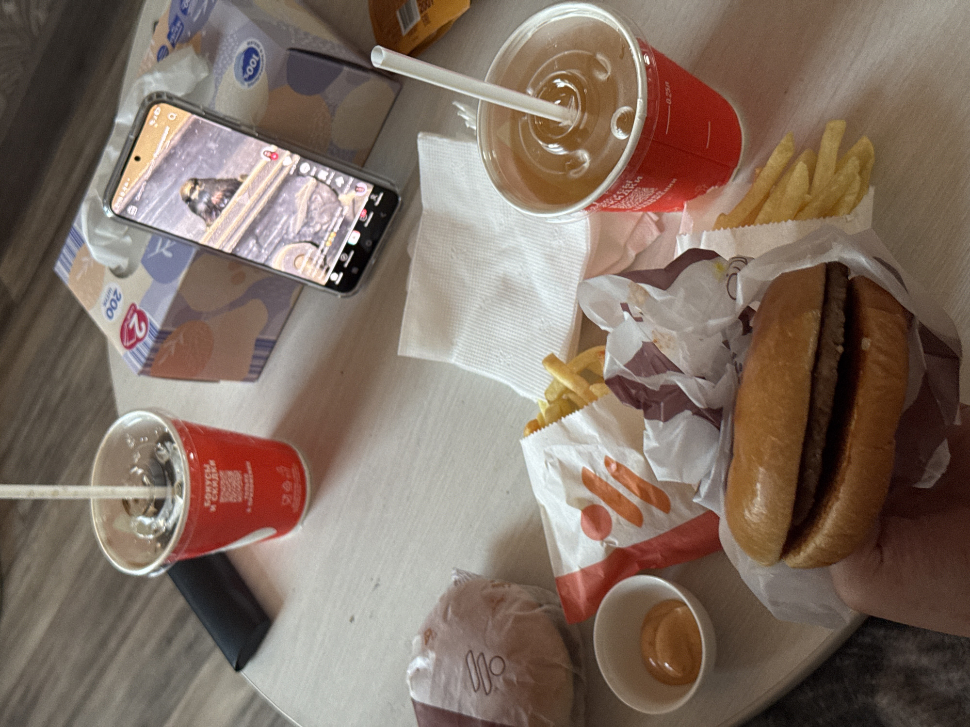 In the city of Zarechnaya (Gamovskoe S/P), Russia, a table is set with fast food and a smartphone. A red cup with a straw and dark liquid, possibly soda, is visible, along with another red cup containing a lighter colored beverage and a straw. A burger wrapped in paper sits in the foreground, held by a hand. Next to it are french fries in a white paper container with orange branding and a small white dish of orange sauce. A smartphone displays a video of a bird on a wooden surface. Two boxes of tissues are positioned behind the phone. The scene suggests a casual meal or break taken indoors.