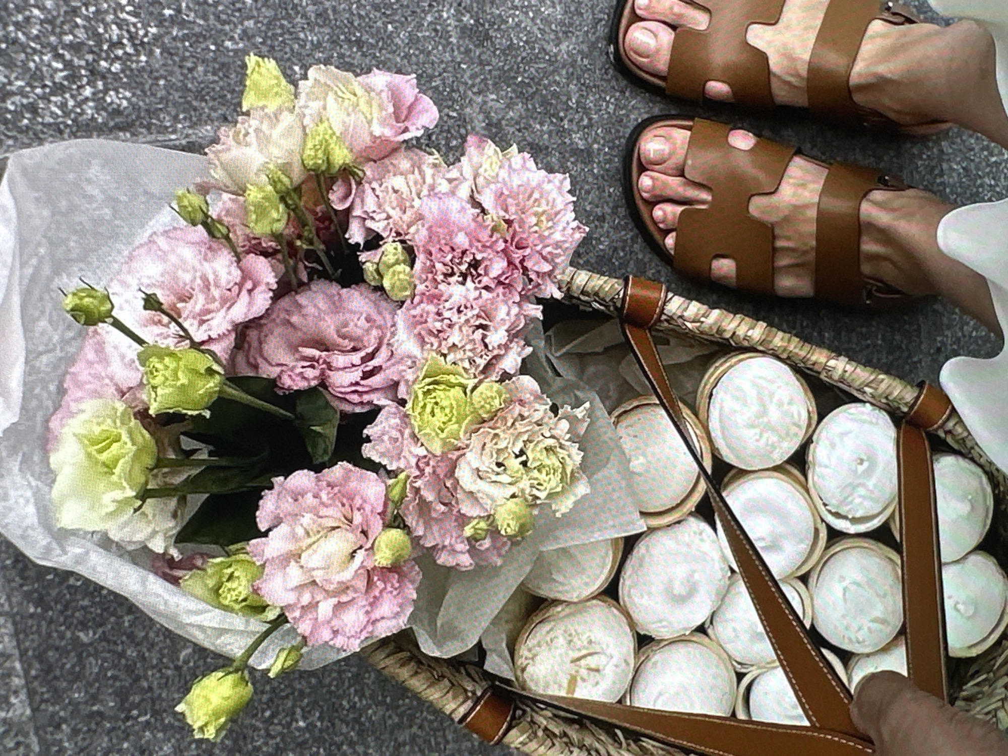 A high-angle shot shows a basket of cupcakes and a bouquet of flowers. The basket is woven with brown leather handles and contains about 12 cupcakes with white frosting. The flowers are light pink and green and are wrapped in white paper. Next to the basket, there is a pair of brown leather sandals with a distinctive "H" design. The feet wearing the sandals are lightly tanned. The ground is gray. The location is not provided in the image.